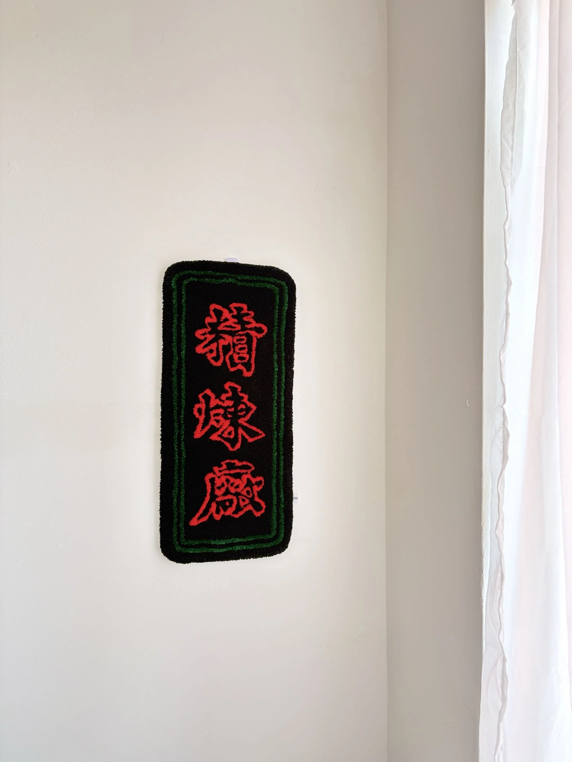 Wall with a black, green, and red decorative hanging ornament featuring Chinese characters, next to a window with white curtains.