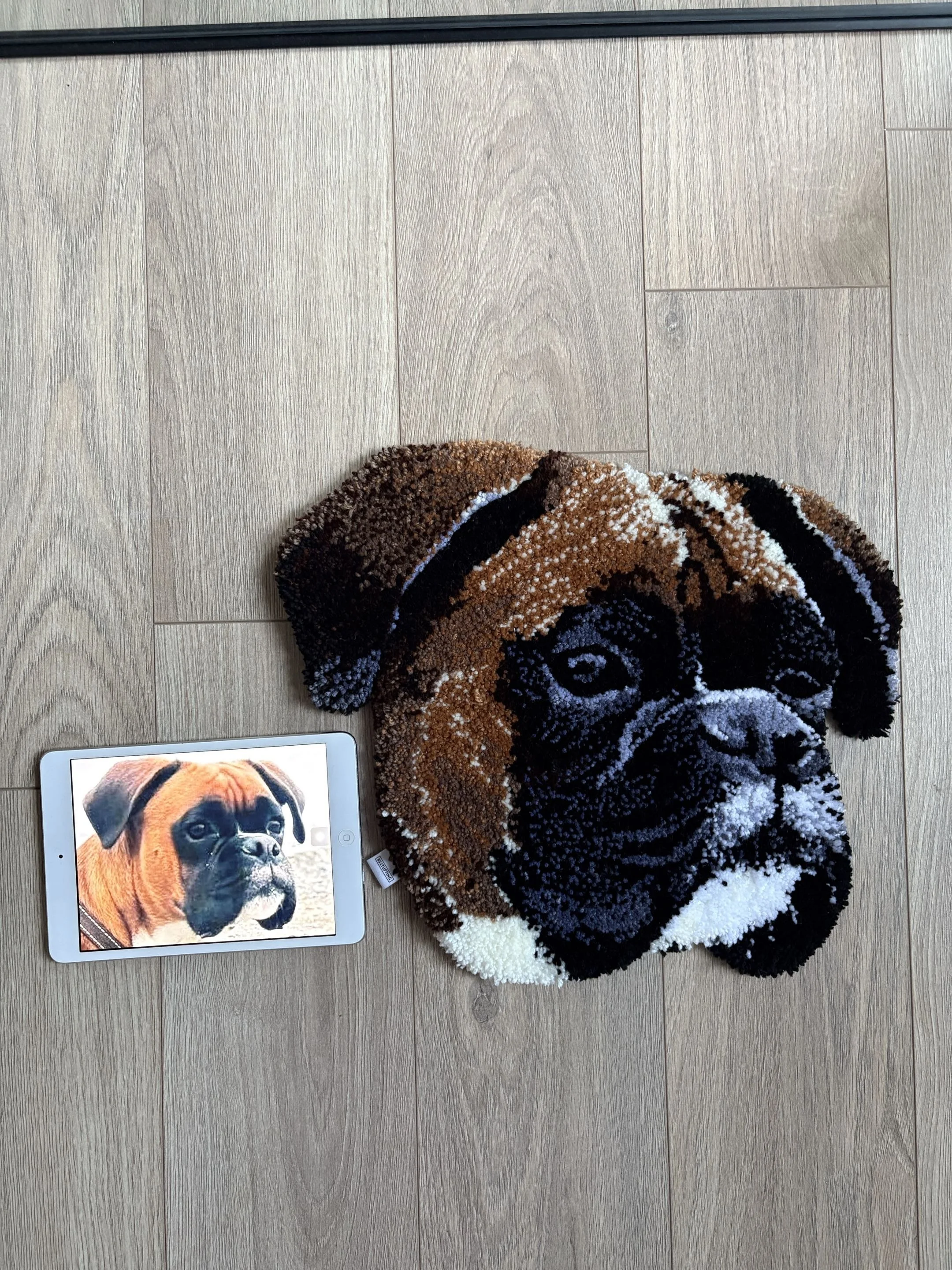 Three circular, plush pet rugs in brown, black, and white with beige, tan, brown, black, and white swirls and patterns, placed on a light wooden floor. A tablet displaying the face of a dog is partially visible at the bottom left corner.