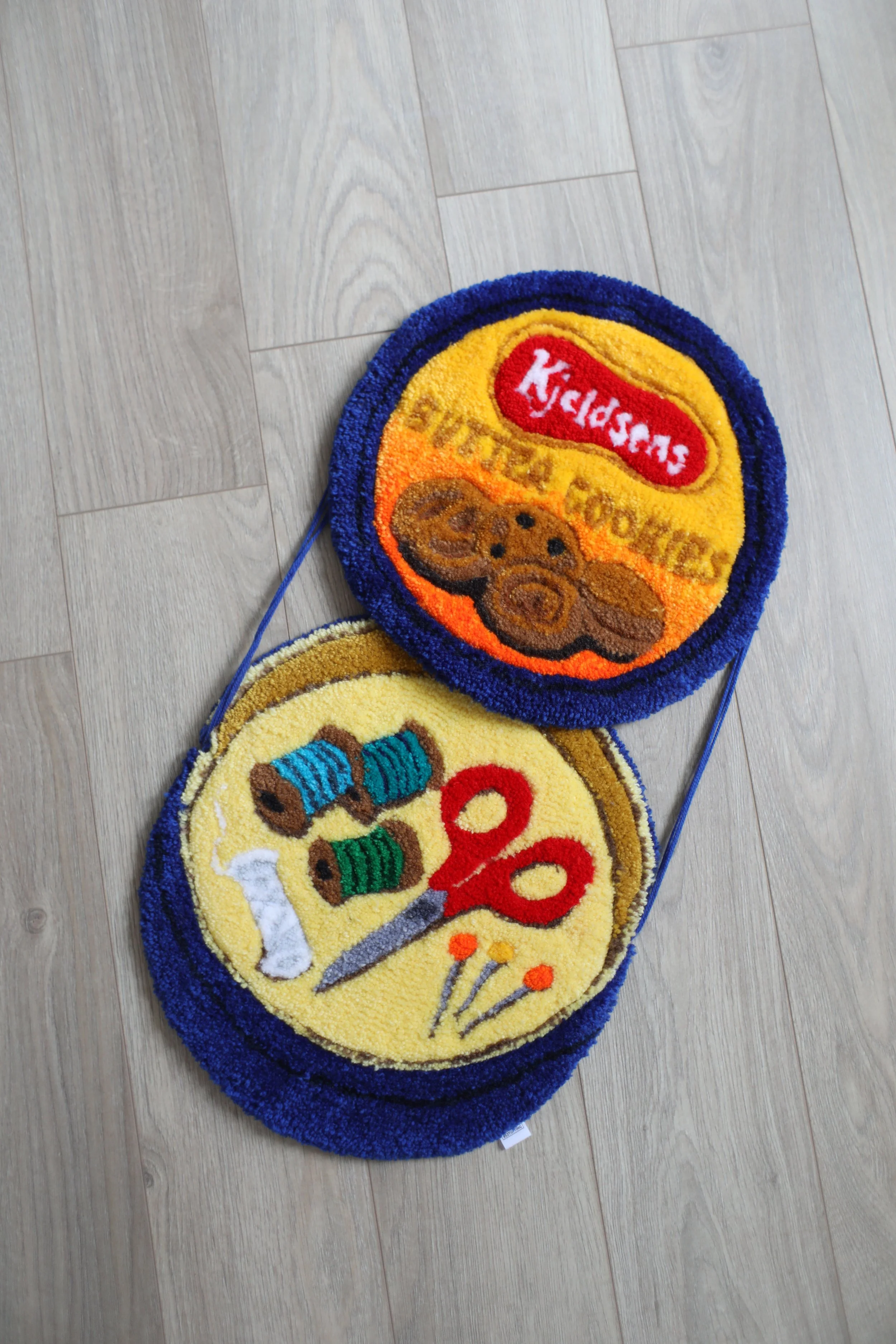Two colorful, oval-shaped doormats on a wooden floor. The top doormat features a brown teddy bear and the words 'Sweet Teddy' in yellow and orange, with a red label that says 'Cookies.' The bottom doormat displays a cupcake with multiple colored topp