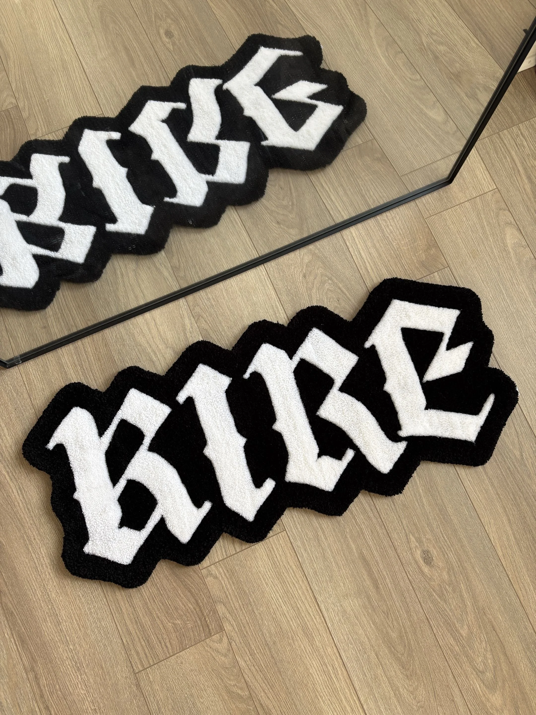 A black and white geometric patterned rug on a hardwood floor, partially reflected in a mirror.