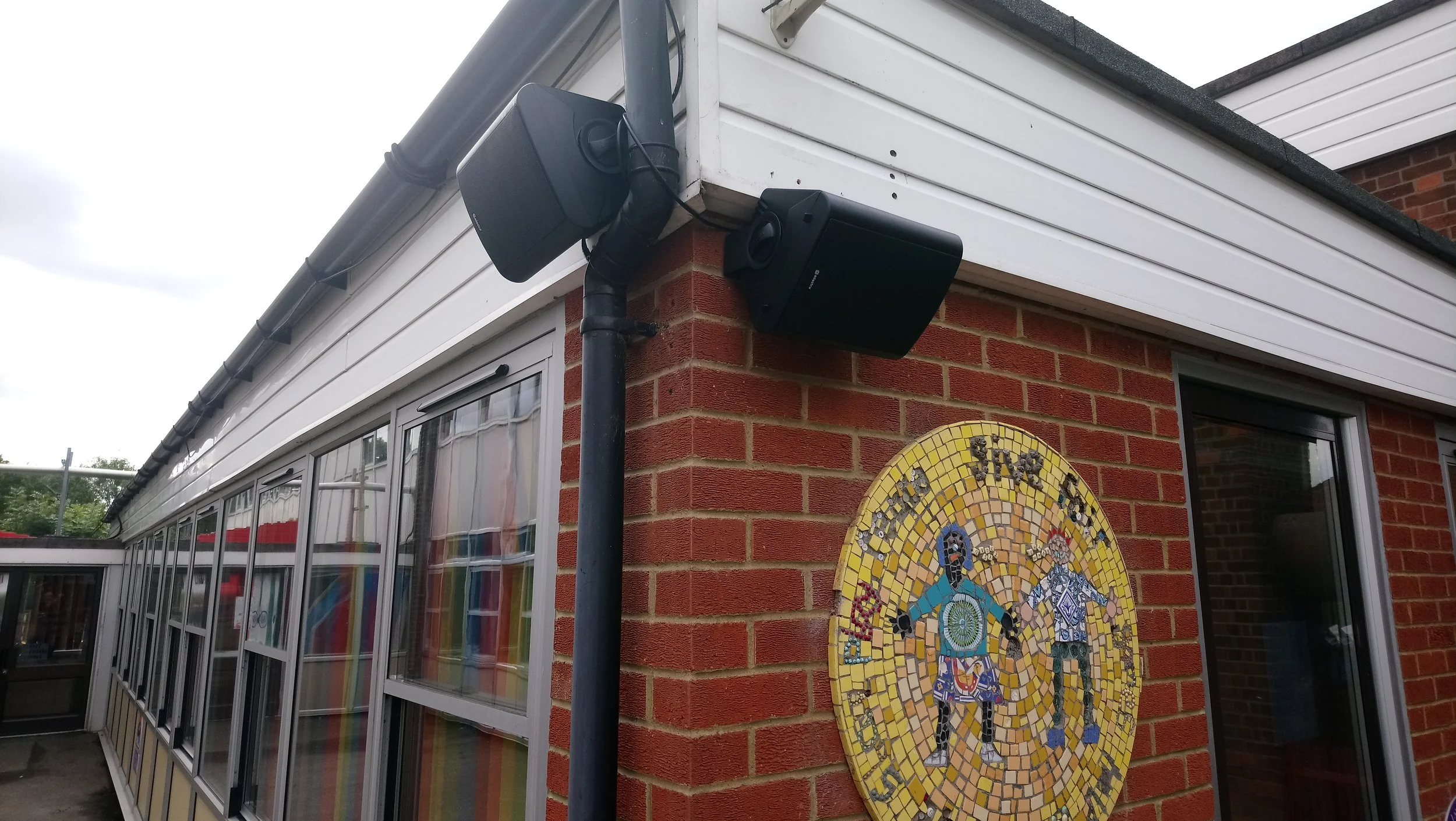 Two black speakers mounted on the exterior of a building, next to a drainpipe, with a colorful mosaic art piece on the brick wall below.