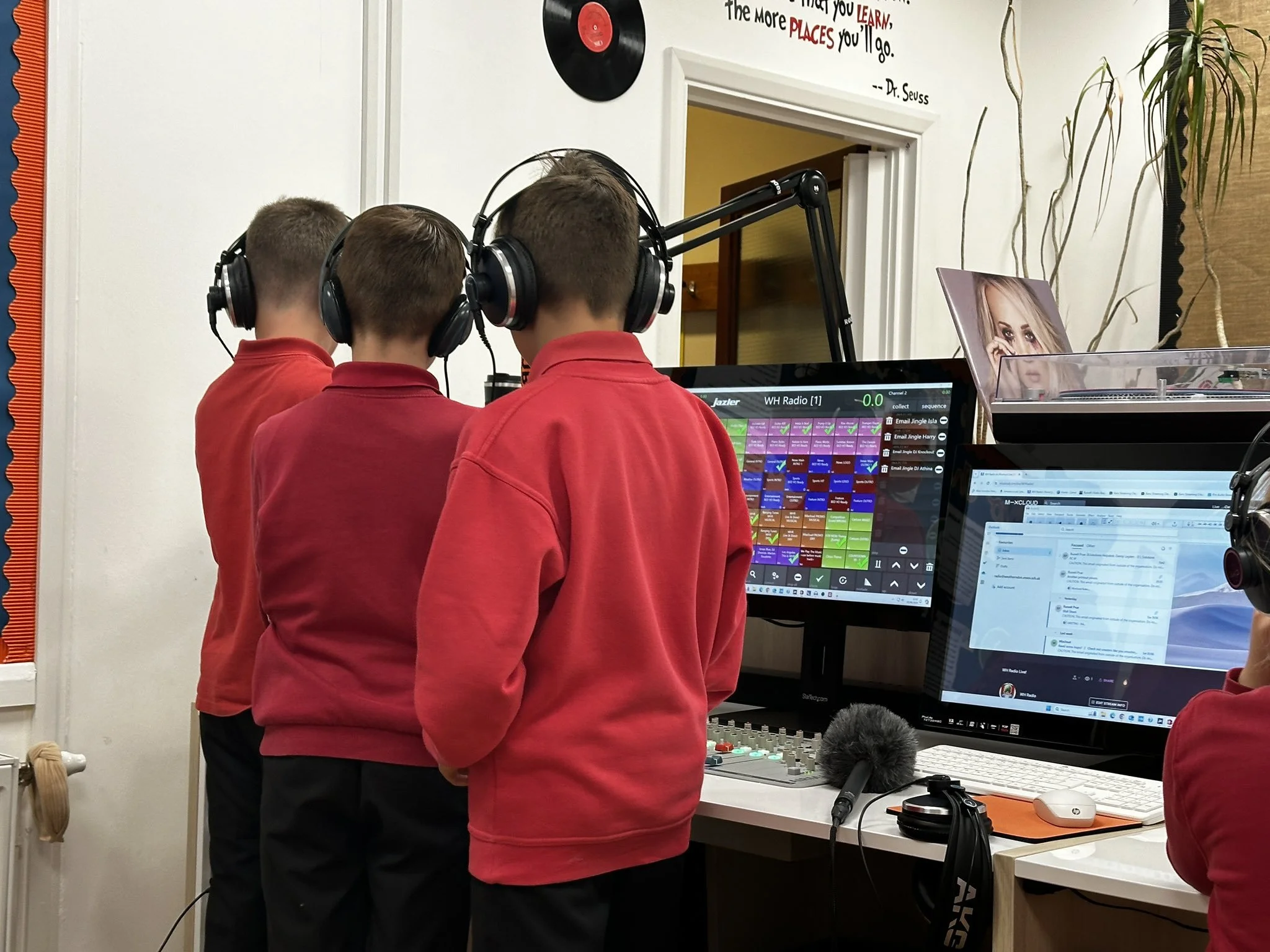 Three students with short hair wearing red shirts and headphones are working on DJing or radio production equipment in a classroom. There are multiple computer screens displaying music or audio editing software, a microphone, and other audio equipment on the desk.