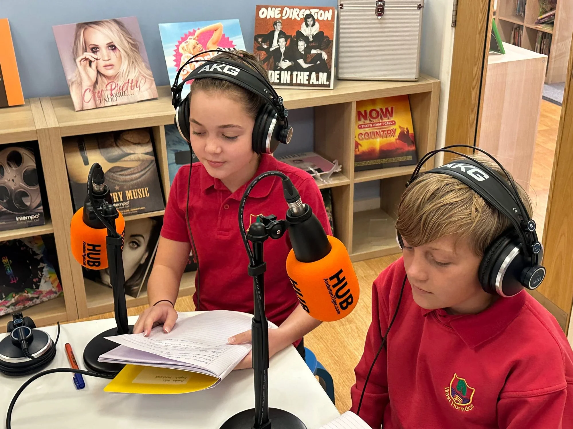 A girl and boy wearing red shirts with a school emblem, sitting at a table with microphones and papers, in a room with music and band posters on shelves behind them.