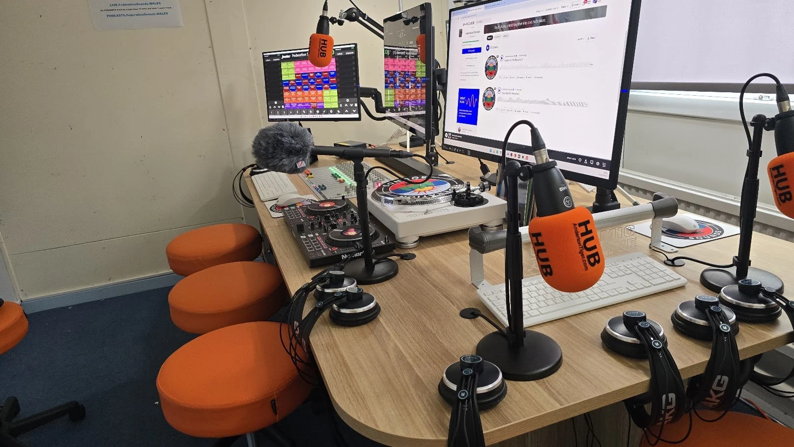 Radio broadcast studio with multiple computer monitors, microphones with orange covers labeled HUB, DJ turntables, a keyboard, and various audio equipment on a wooden table surrounded by orange stools.