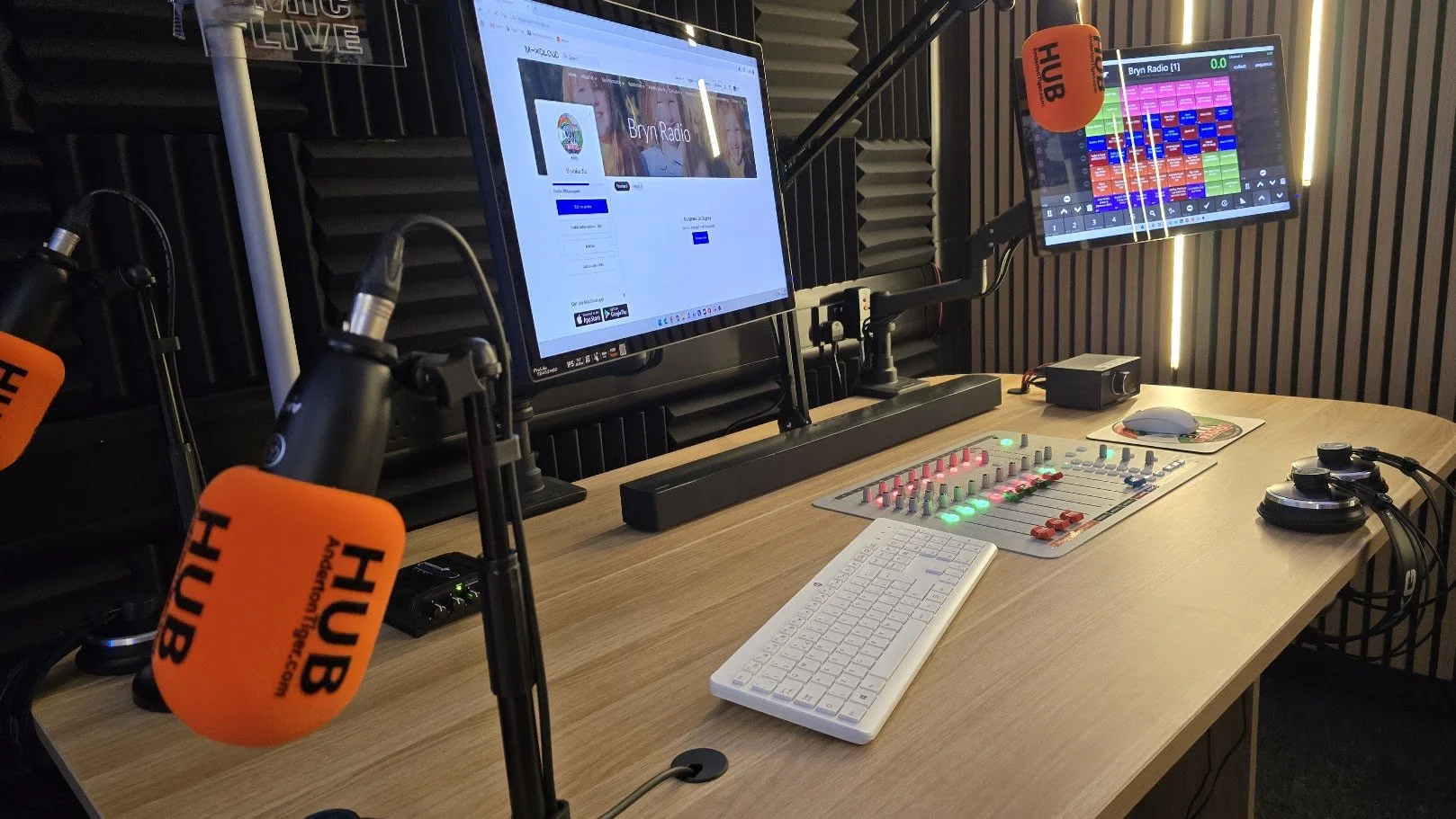 Radio broadcast studio with two microphones labeled HUB, two computer monitors, a sound mixer, a keyboard, headphones, a mouse, and a small audio interface on a wooden desk.
