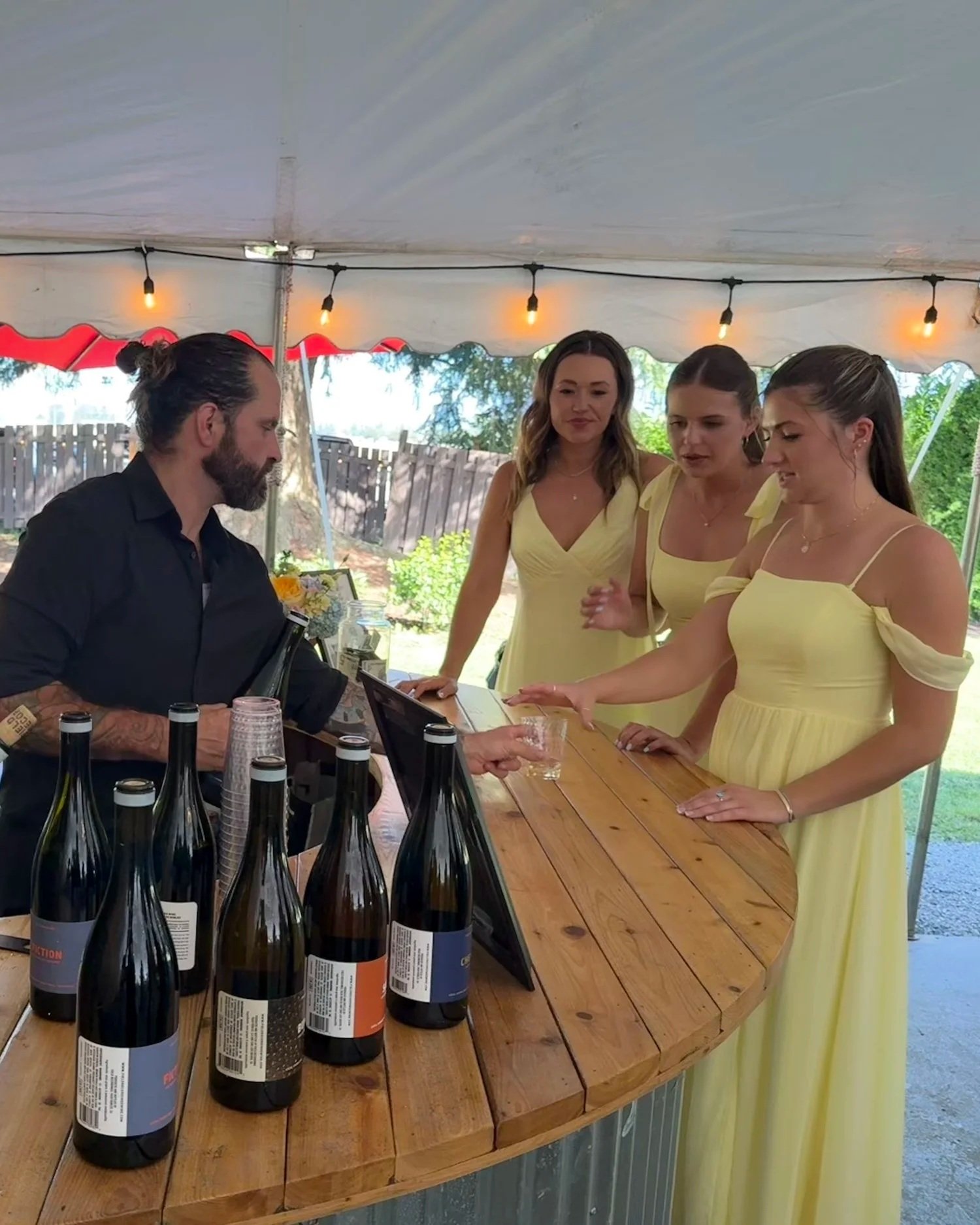People at a wine tasting booth inside a white tent, with bottles of wine on the table and a person pouring wine into a glass.
