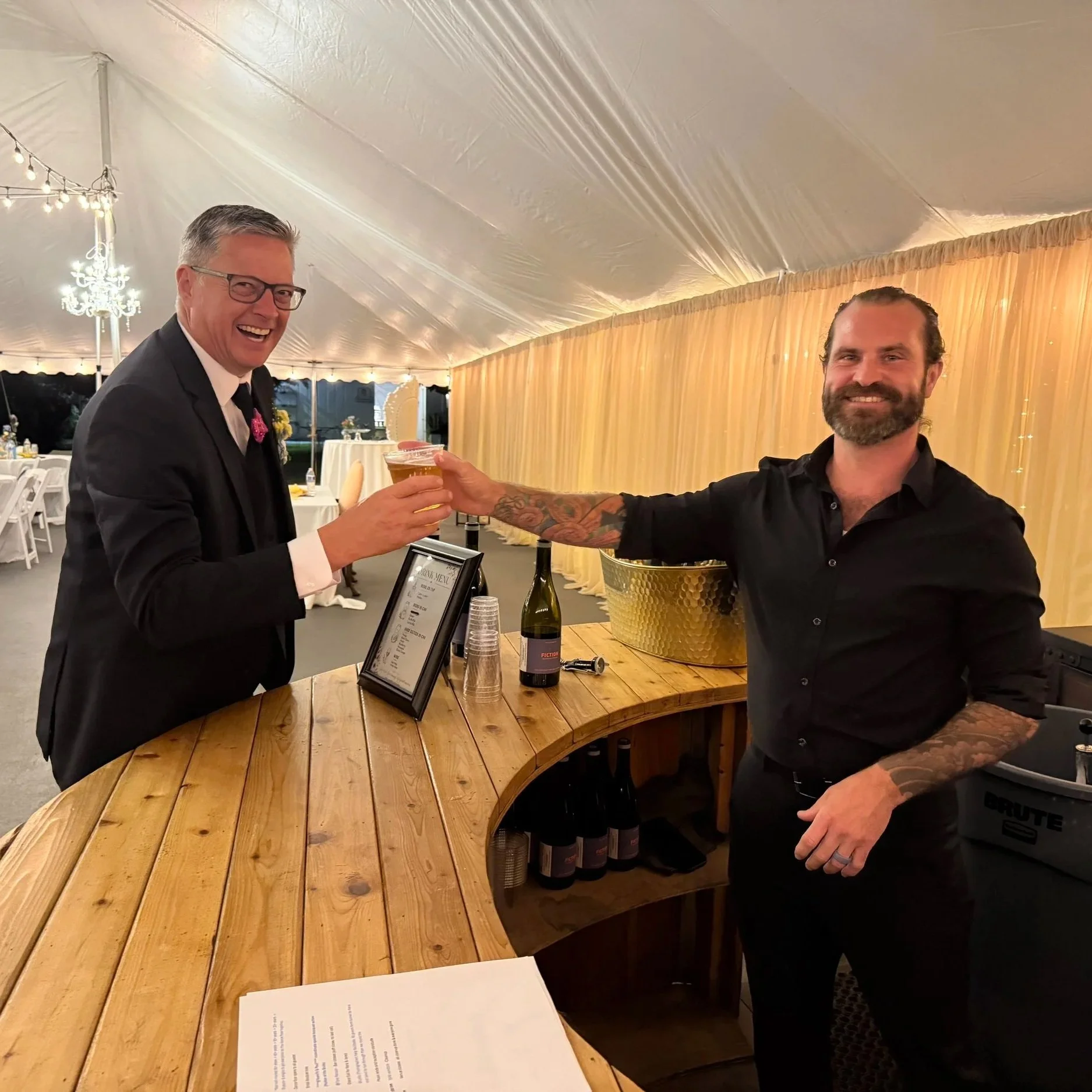 Two men smiling and clinking glasses of beer at a reception under a large white tent, with string lights and draped curtains in the background, and a curved wooden bar with bottles and cups.