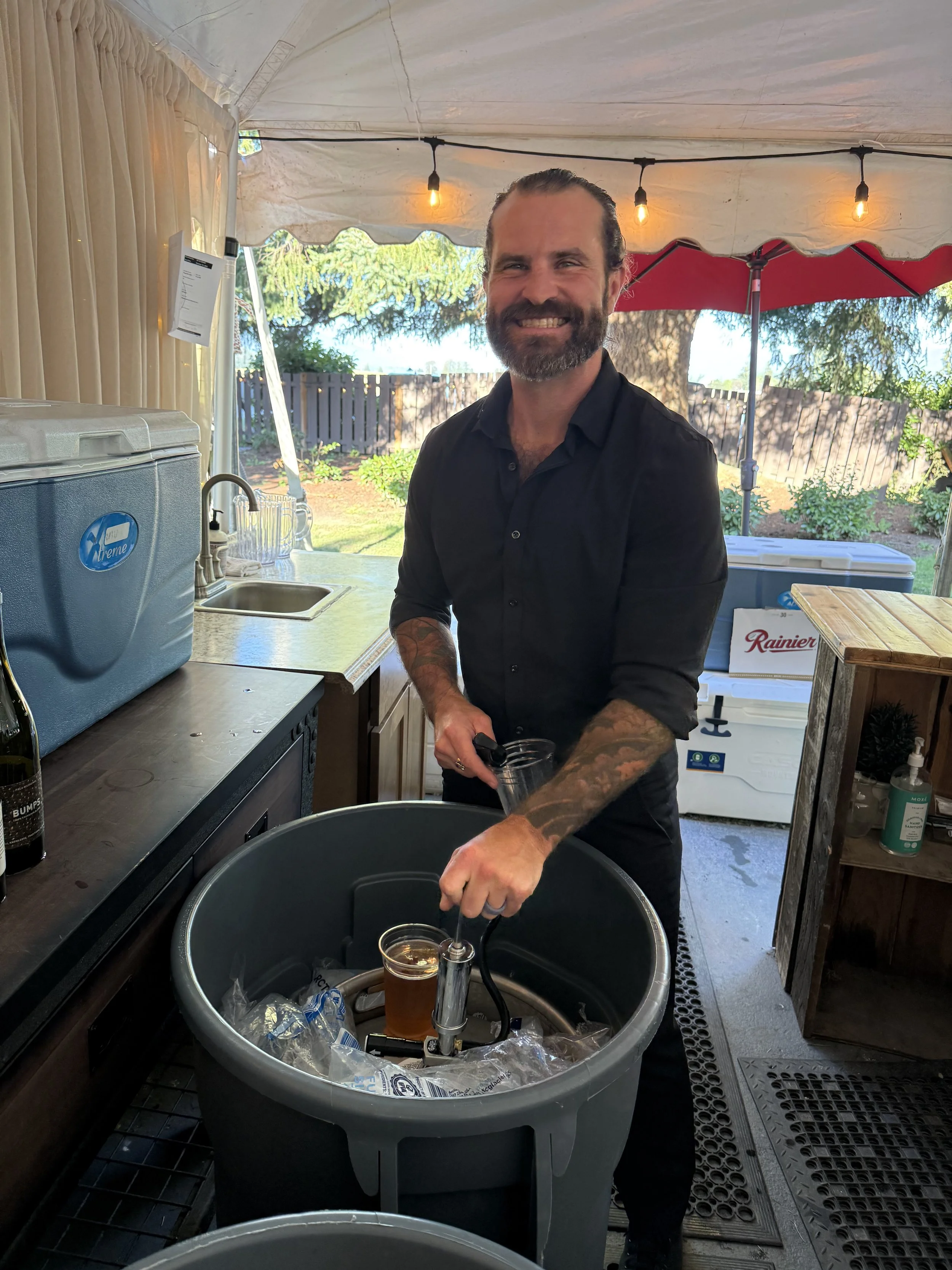 A man with a beard and tattoos on his arms is smiling and pouring liquid into a container inside a tent with string lights. There are coolers, plants, and trees in the background.