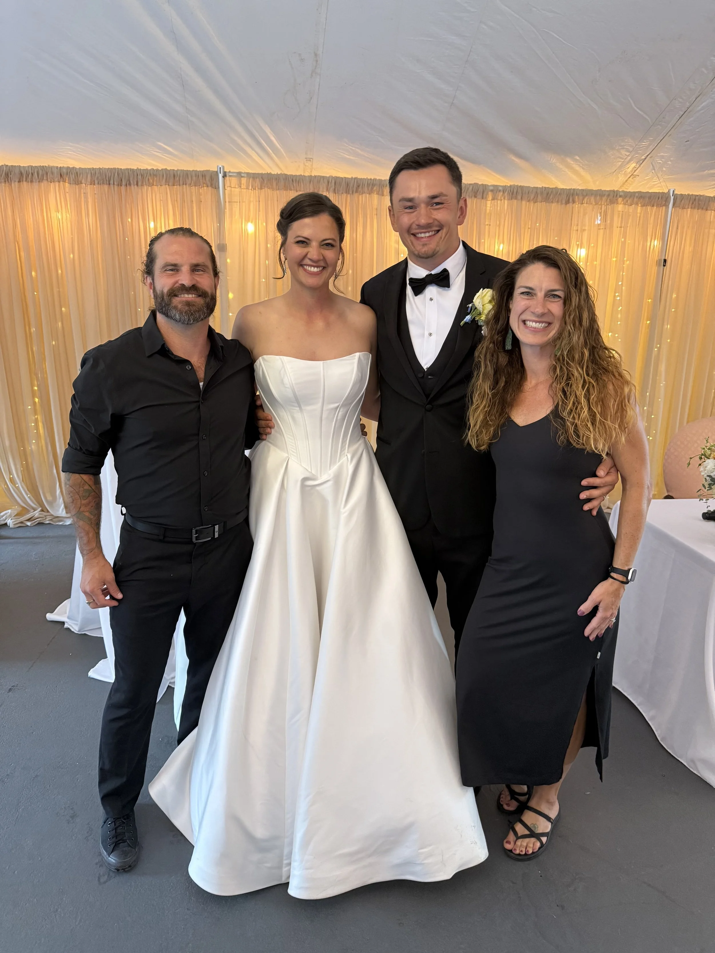 Four people standing together at a wedding reception under a decorated tent, smiling at the camera.