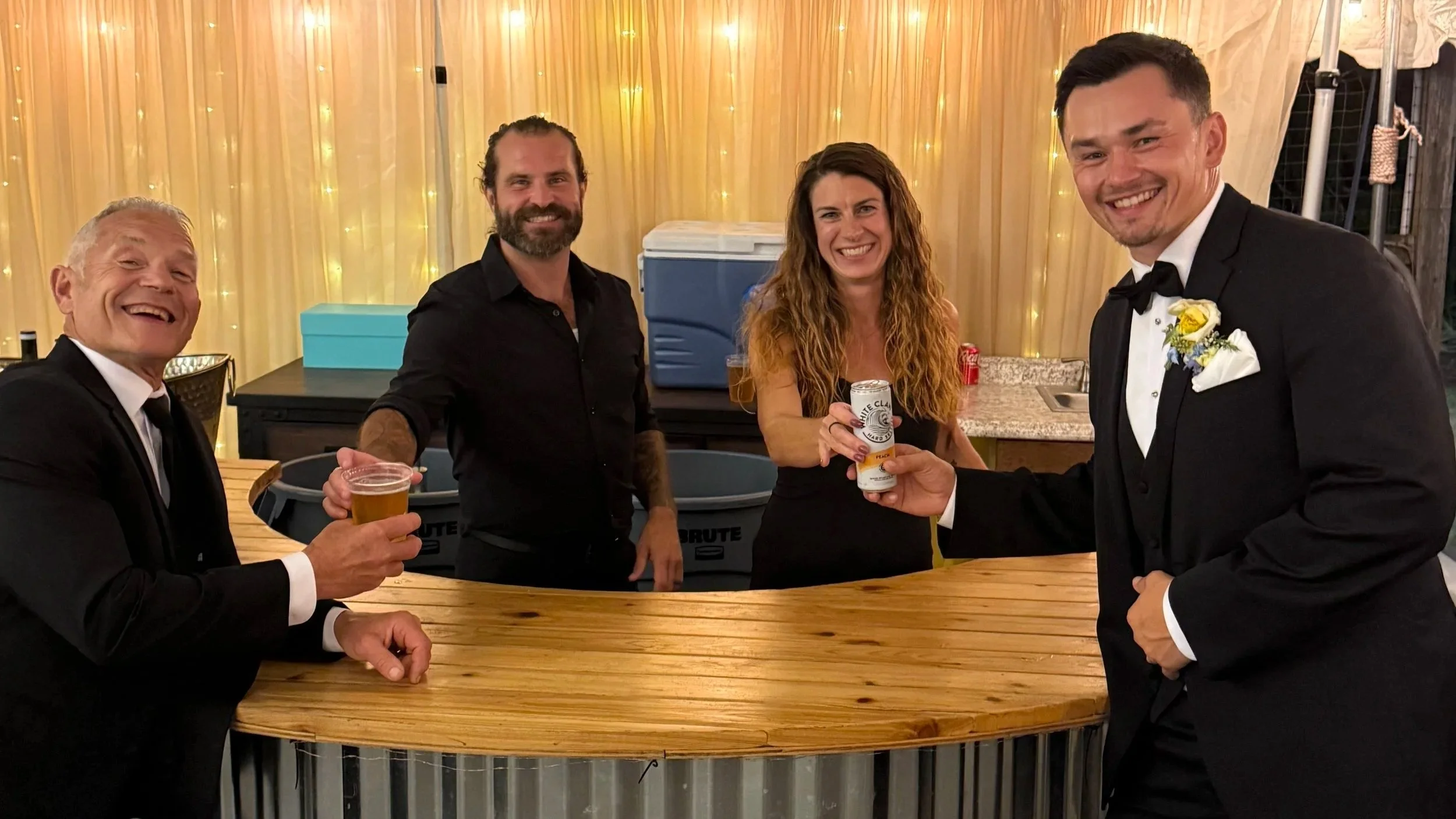 Group of five people dressed in formal attire at a celebration, with three men and two women smiling and holding drinks, indoor setting with yellow curtains and string lights in the background.