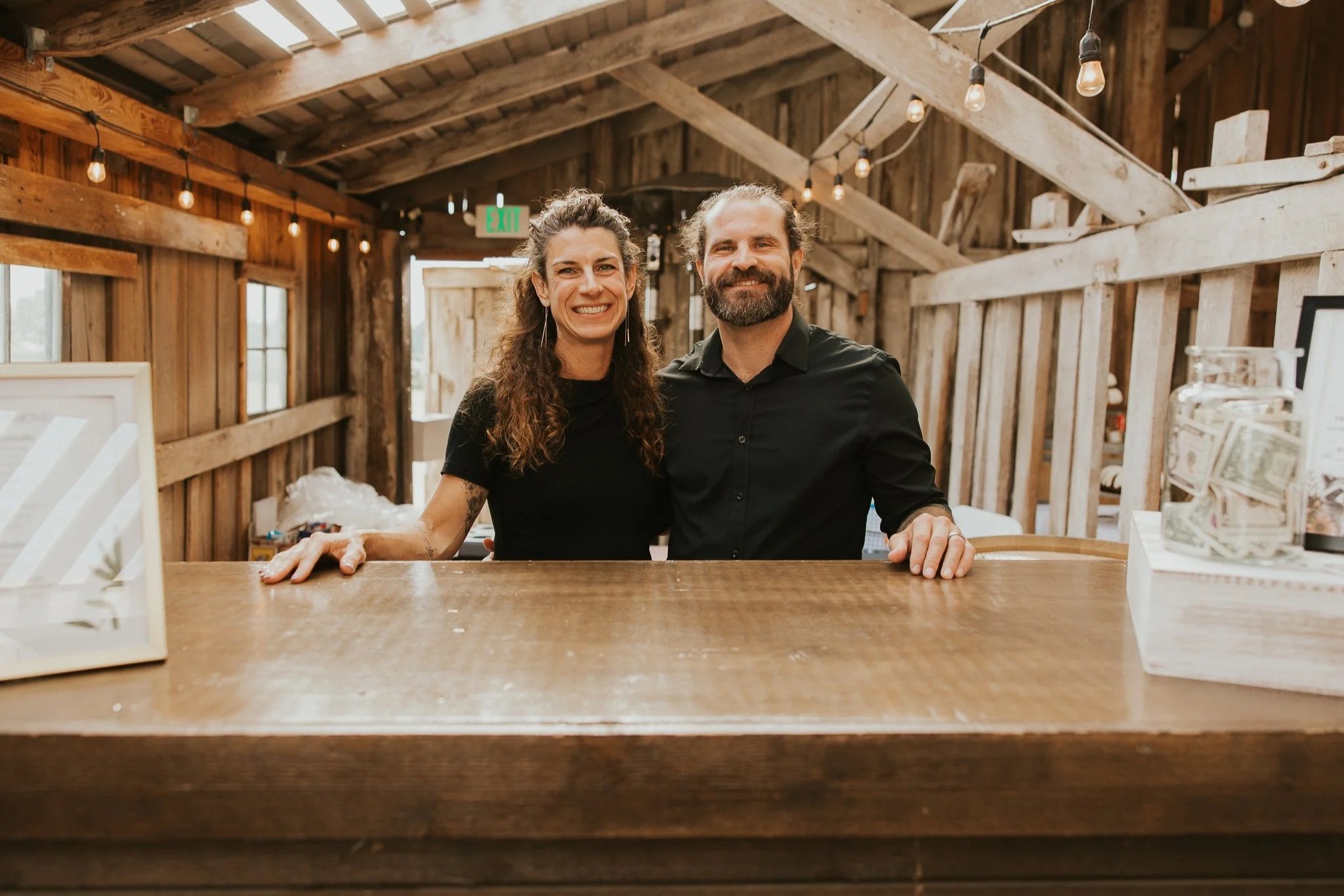 A smiling man and woman standing behind a bar or counter in a rustic wooden building, with exposed wooden beams and hanging light bulbs.