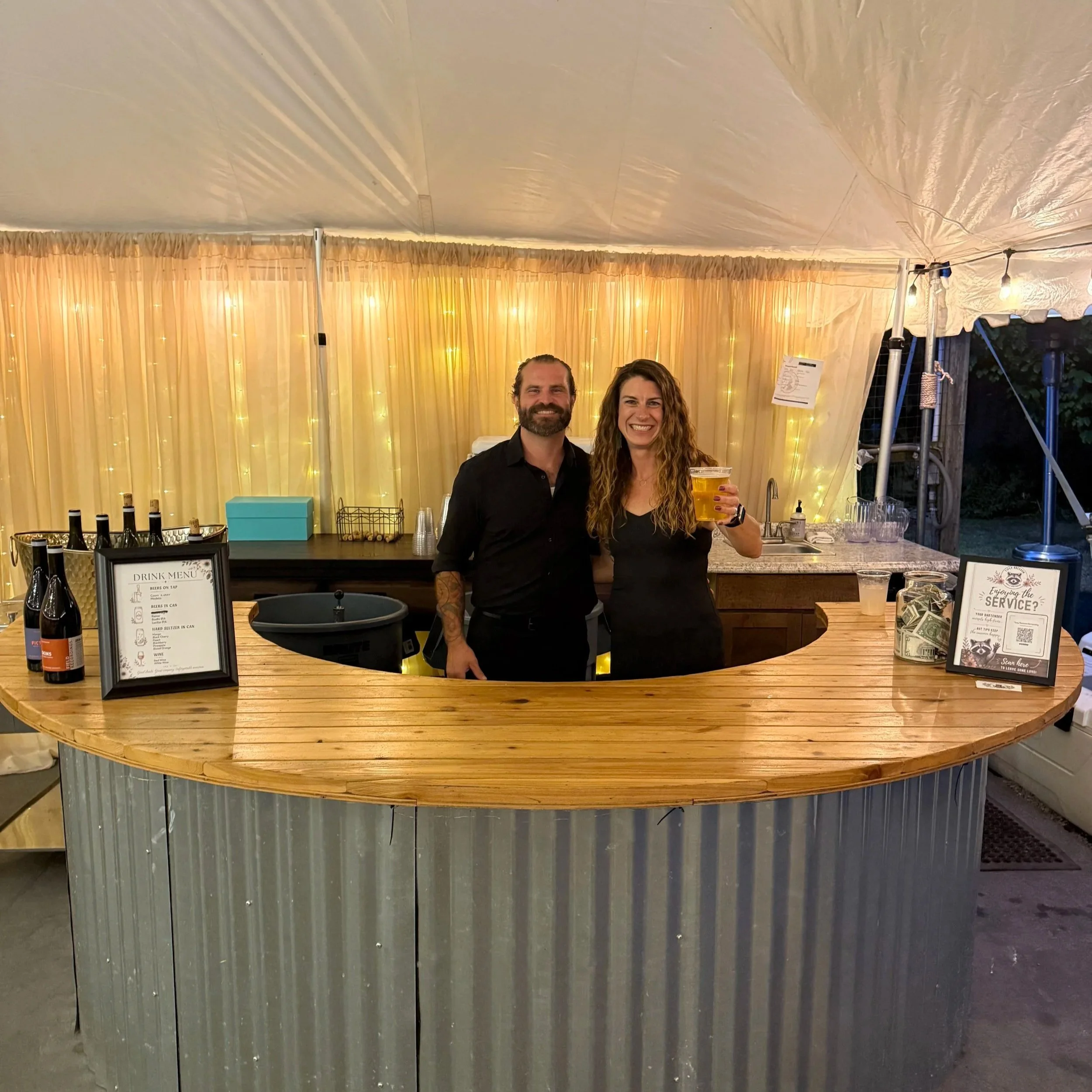 People standing inside a decorated tent at night, with string lights and a bar counter with bottles and menus.