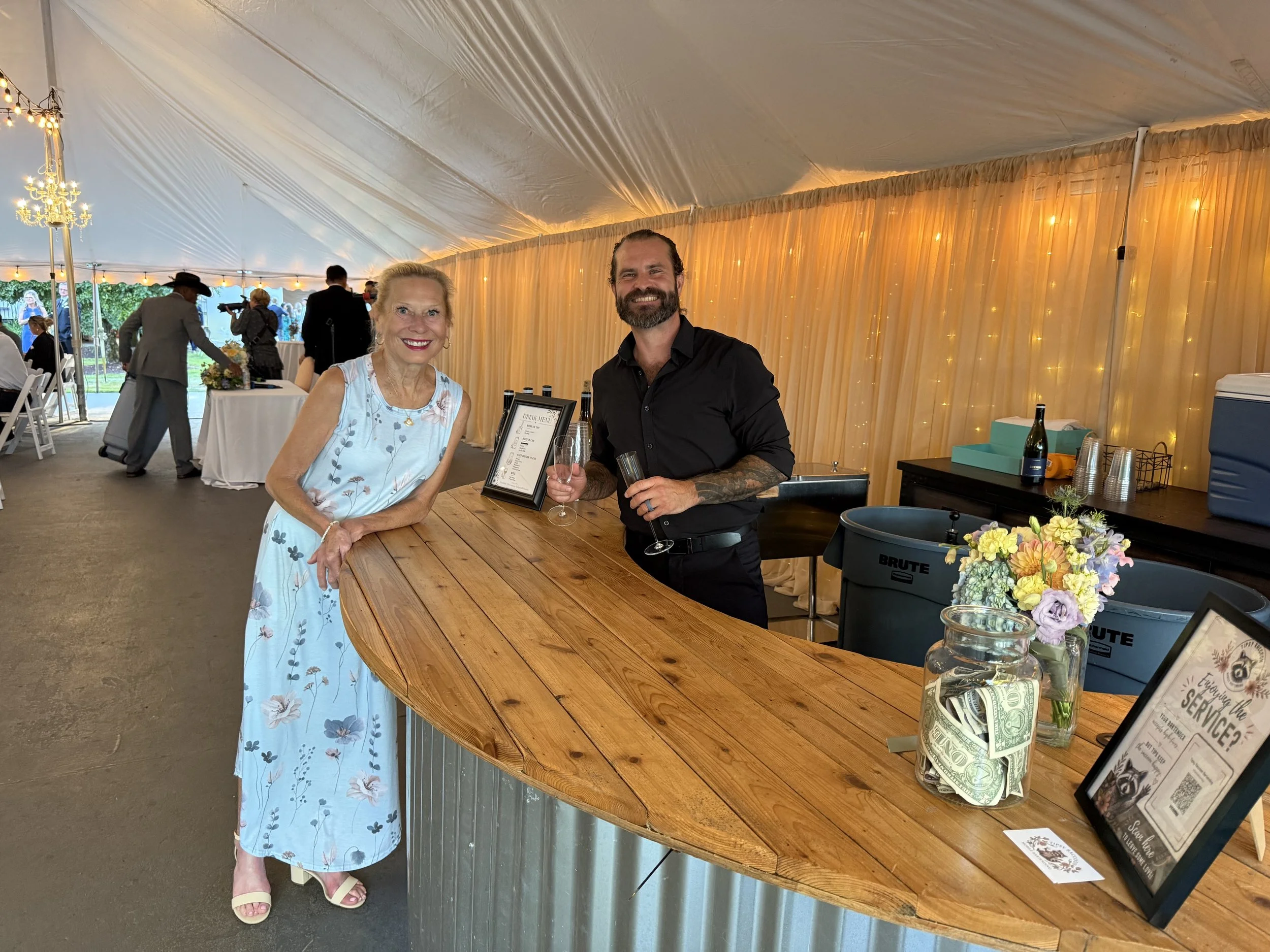 A woman in a light blue floral dress and a man with a beard in a black shirt, smiling and holding glasses of wine, standing at a wooden bar inside a decorated event tent with warm lighting and gold-colored curtains.