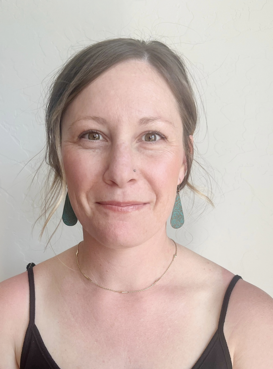 A young woman smiling with short brown hair, wearing a black tank top, turquoise earrings, a delicate gold necklace, standing against a plain white wall.