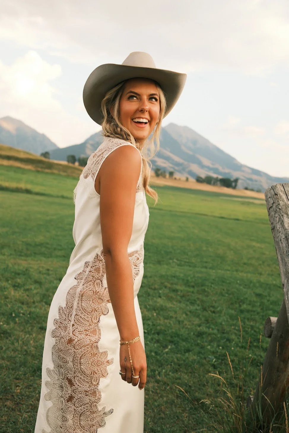 A smiling woman wearing a wide-brimmed hat and a sleeveless lace dress standing in a grassy field with mountains in the background.