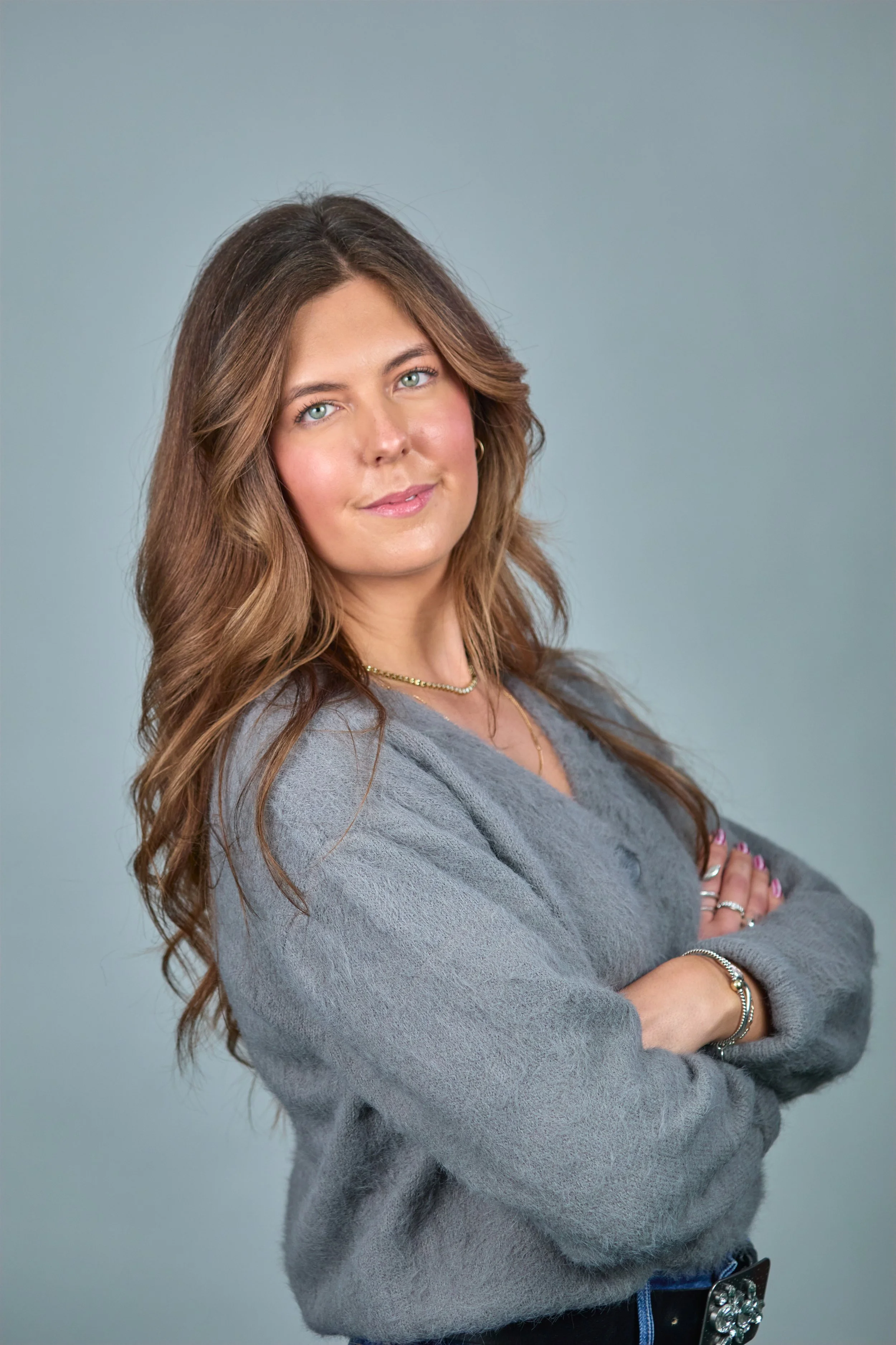 A woman with long, wavy brown hair and green eyes, wearing a gray sweater, jewelry, and with arms crossed, posing against a plain gray background.