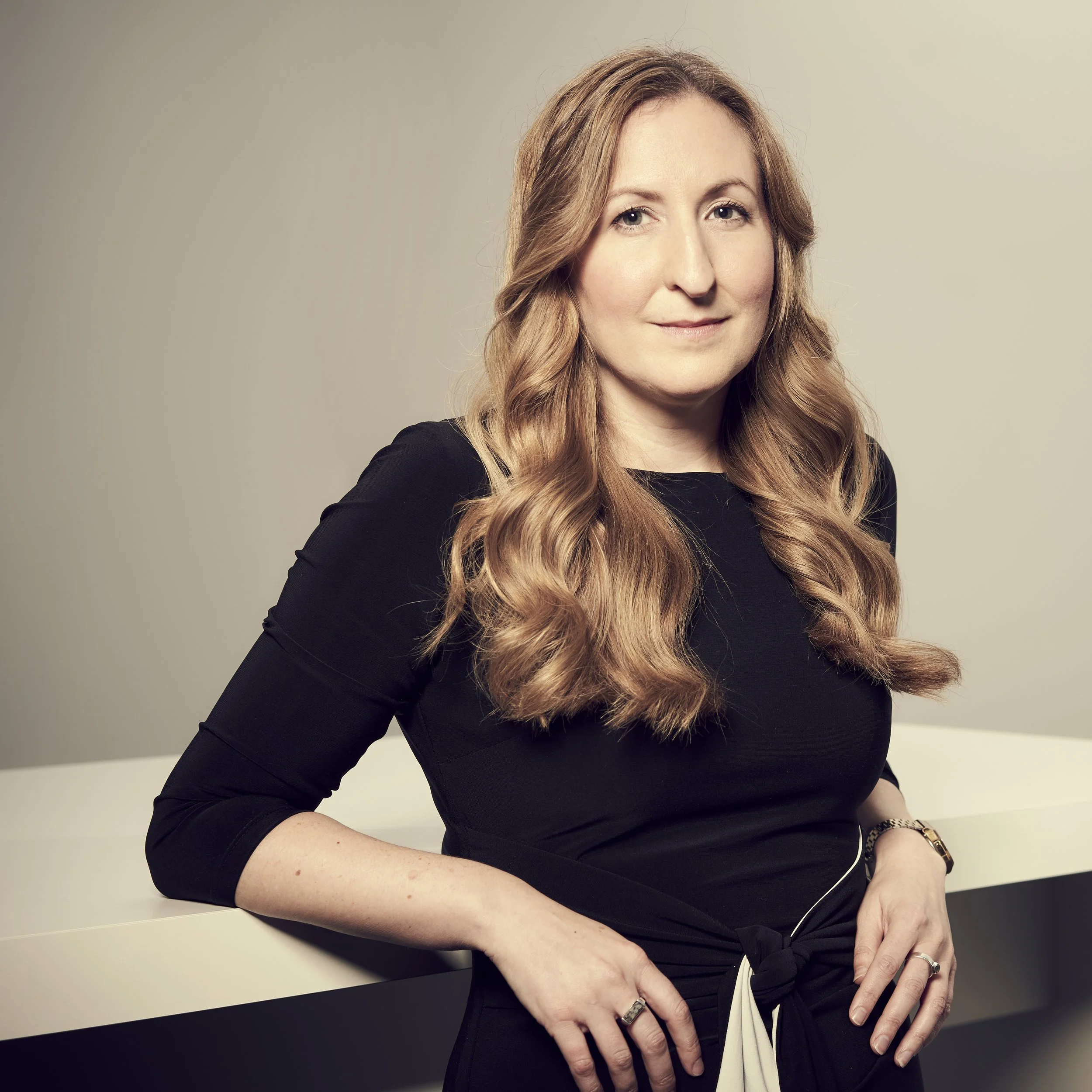 Close-up of a woman with long fair hair wearing a black top, sitting at a table against a plain light background. She has a subtle smile, light makeup, and wears multiple rings and a watch.