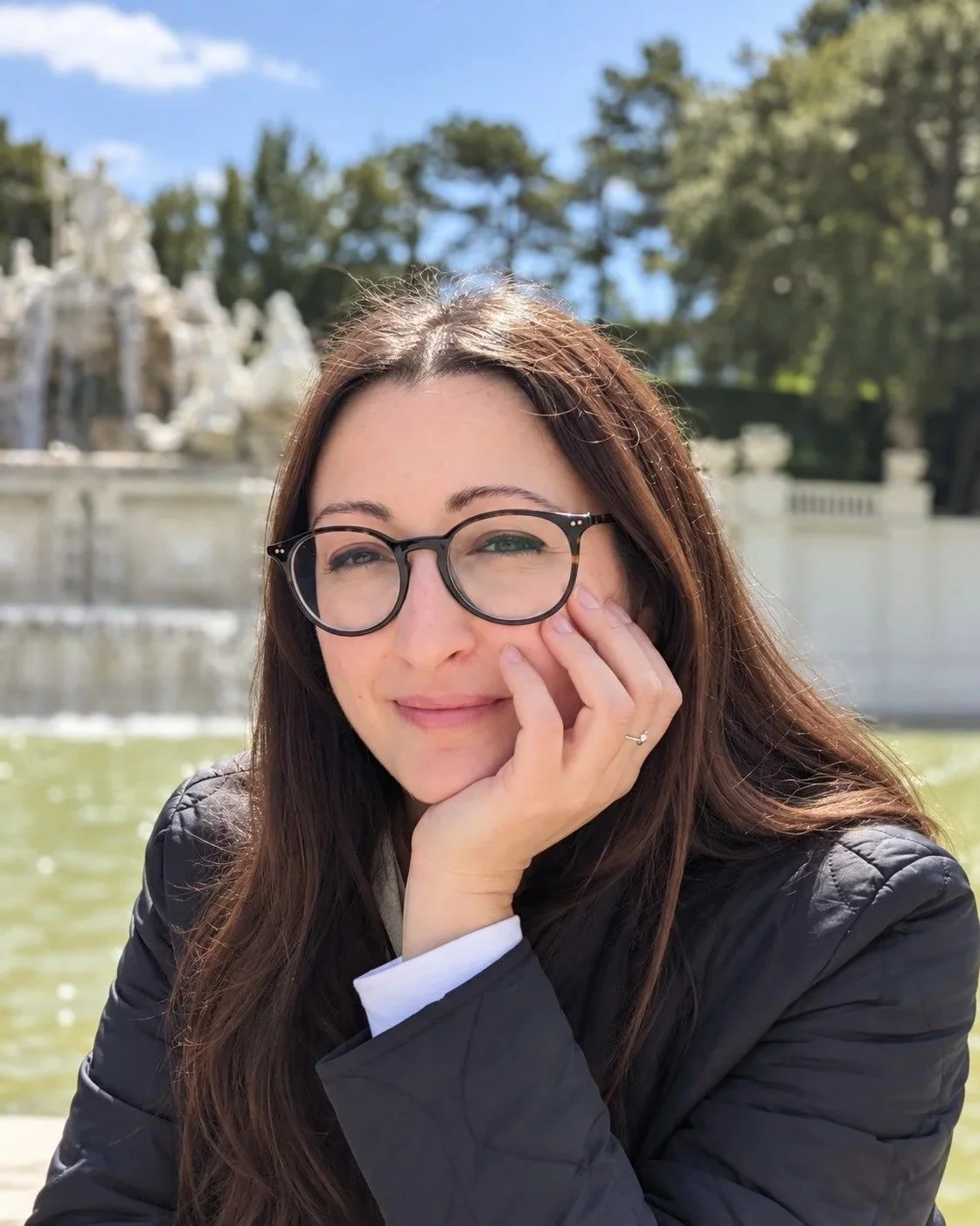 A woman with long brown hair and black glasses sitting outdoors near a fountain, with trees and a stone structure in the background on a sunny day.