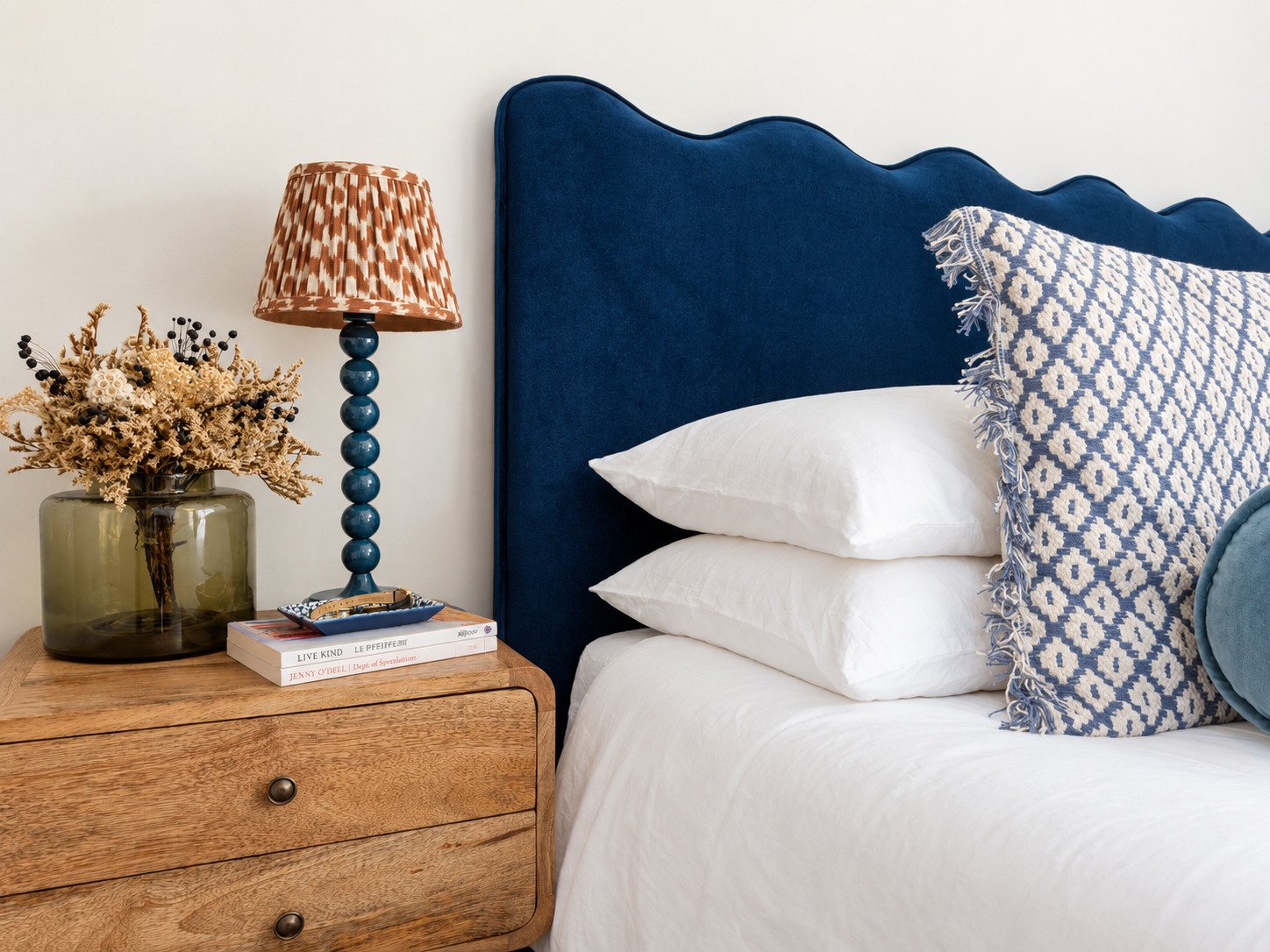 A cozy bedroom setup with a wooden nightstand, a green vase with dried flowers, a stack of books, a blue table lamp, a bed with a blue upholstered headboard, white pillows, and a patterned pillow.