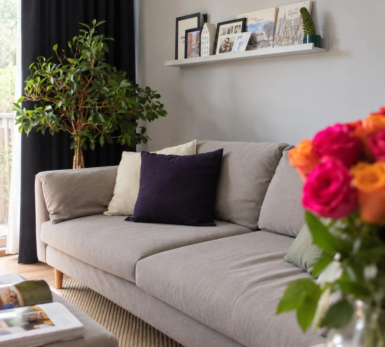 Living room with a beige sofa adorned with purple, cream, and green pillows, a large potted plant, a white wall shelf with photos and decor, and a bouquet of pink and orange flowers in the foreground.