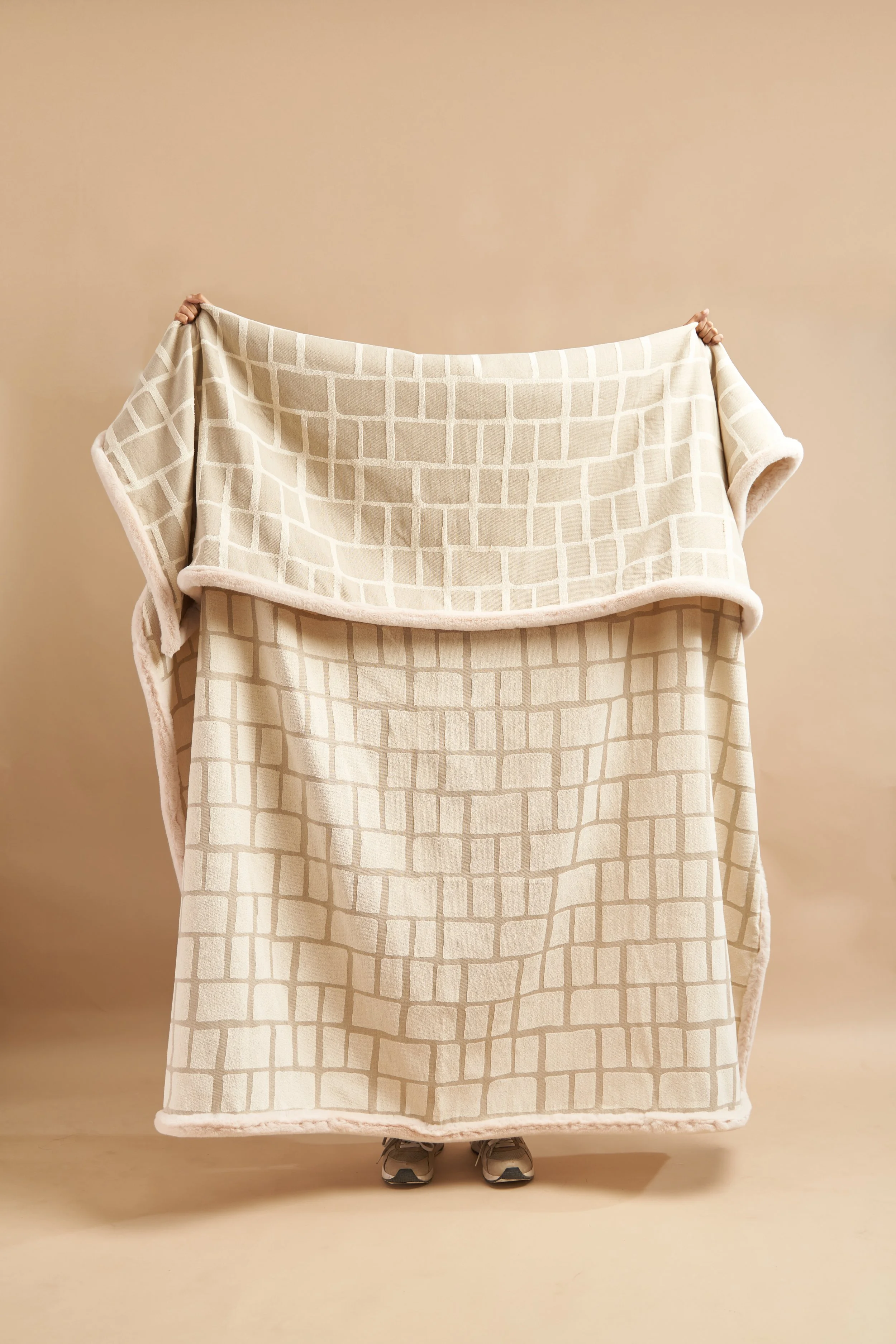 Person standing behind a large beige blanket with a geometric pattern, holding it up. Only the person's shoes and part of their hands are visible.