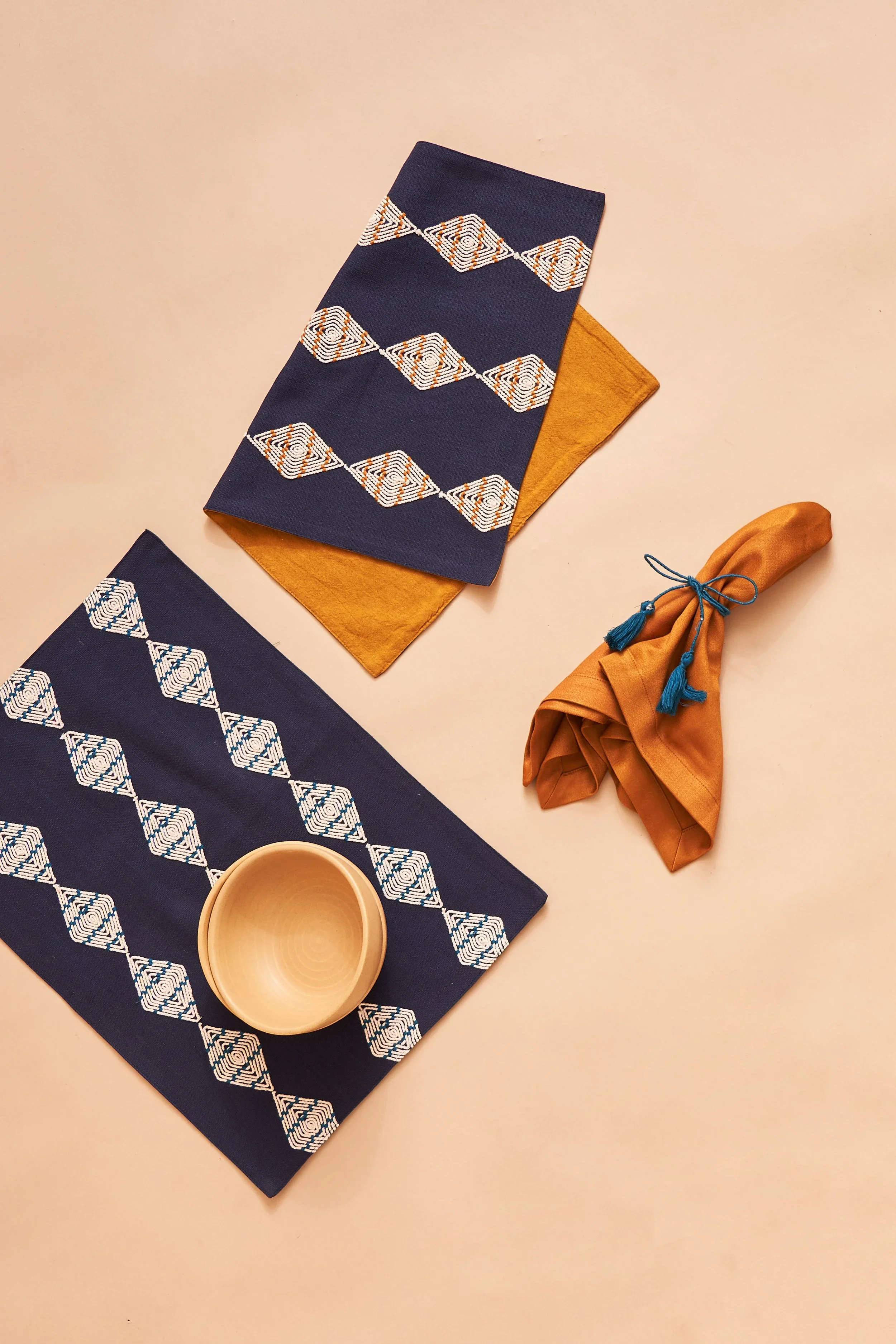 Table setting with a navy blue placemat featuring geometric white and orange embroidery, a mustard yellow napkin with blue string, and a beige ceramic bowl on a light-colored table.