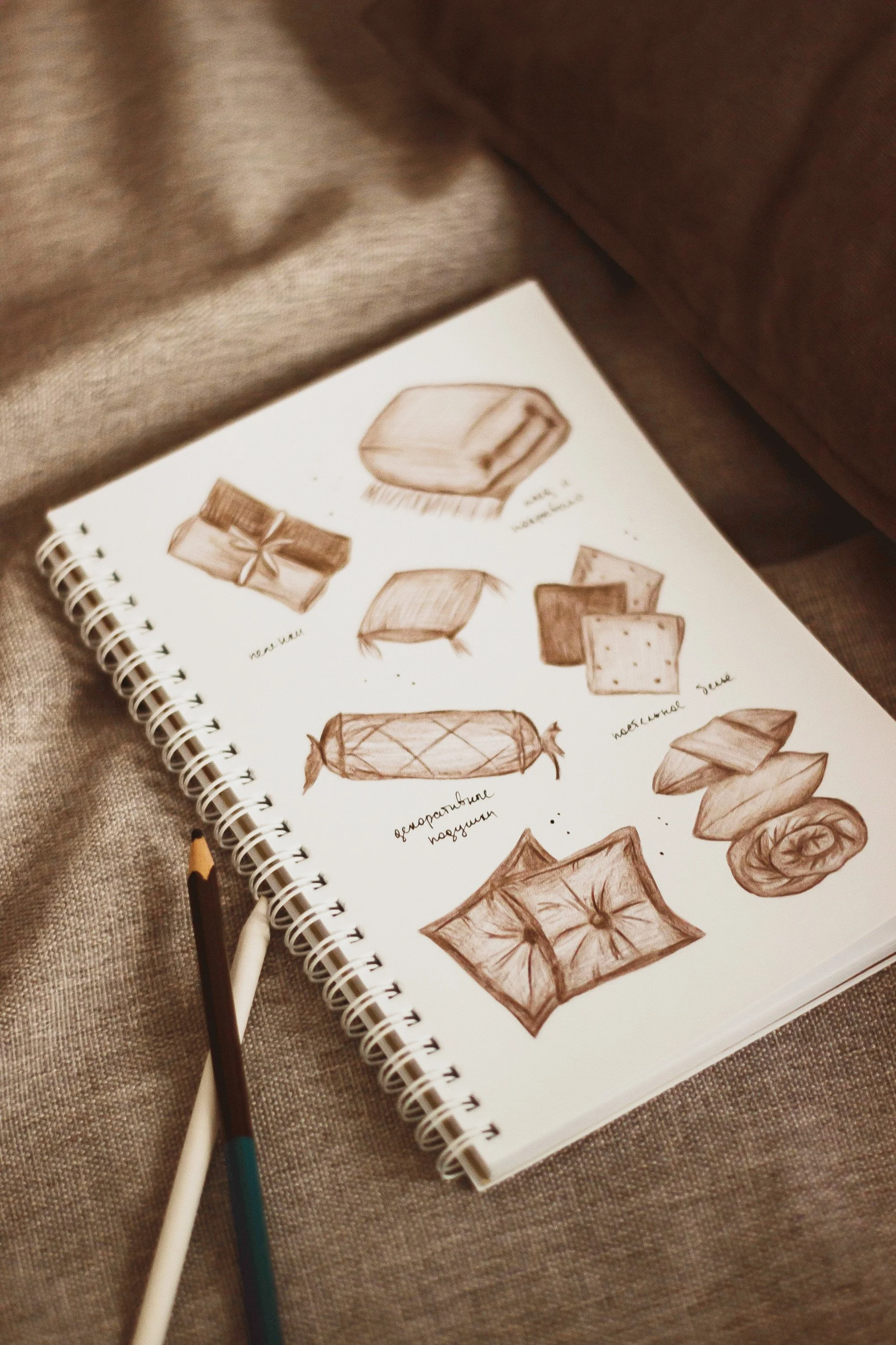 Sketches of various sofa and furniture designs drawn in brown ink on a spiral notebook page, resting on a beige fabric surface with two pens nearby.