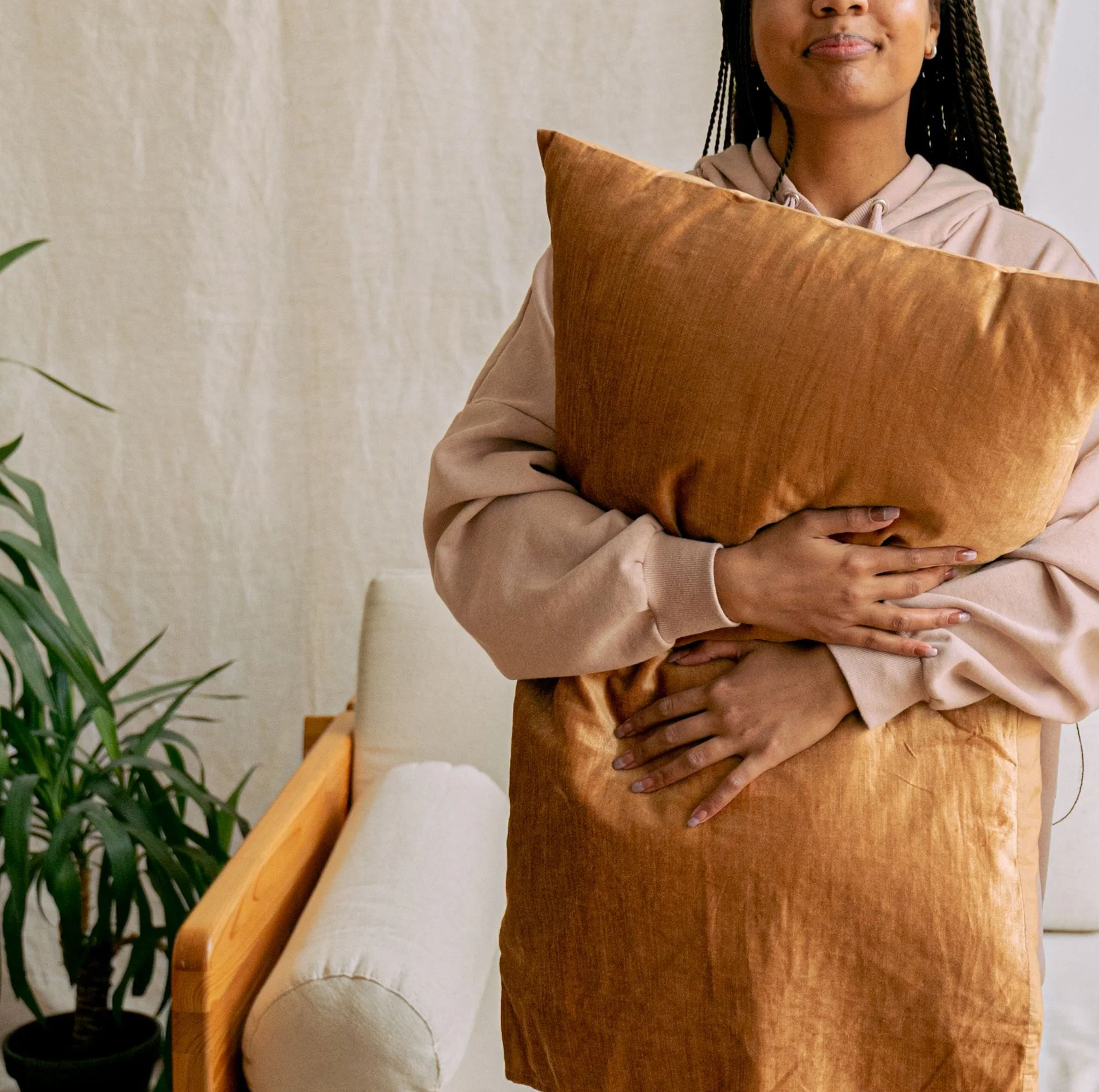 Person holding an orange pillow, wearing a beige hoodie, standing near a wooden bench and green plant in a room with white walls.