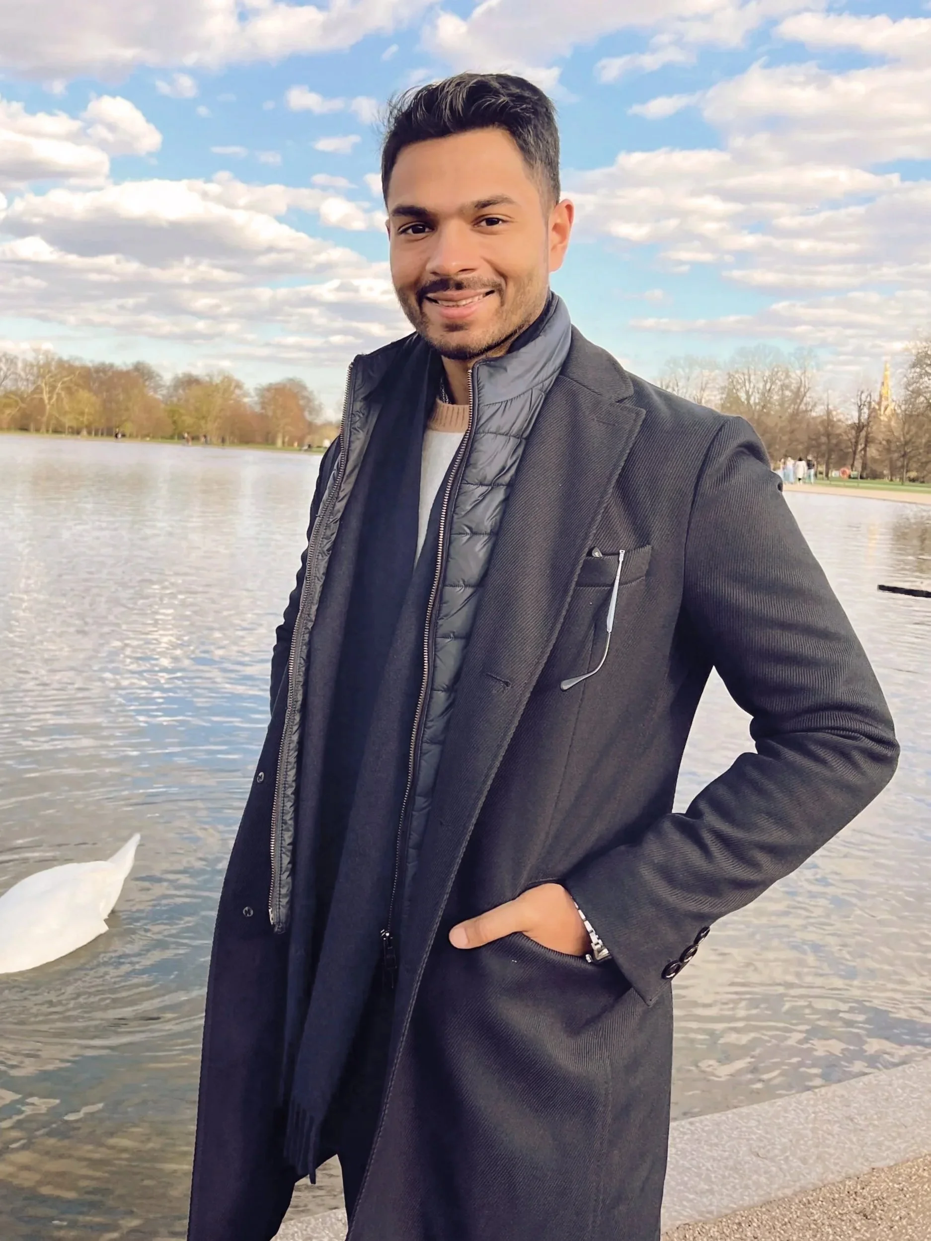 A man in a black coat and gray vest standing by a body of water with trees and a cloudy sky in the background.