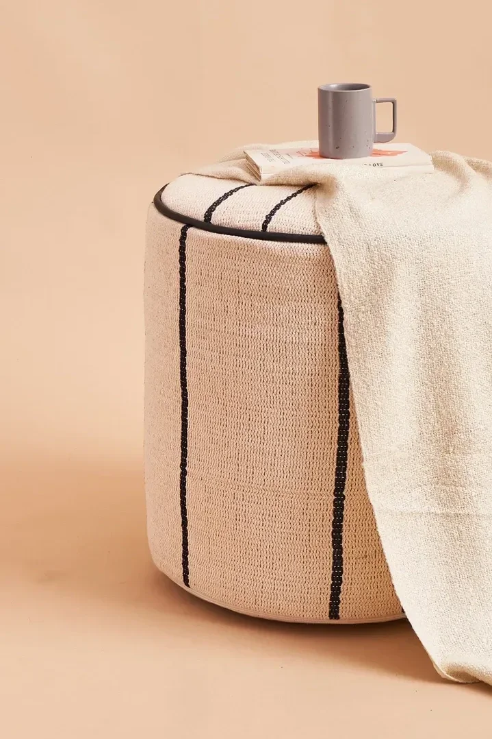 A round beige fabric storage ottoman with black stripes, topped with a white textured blanket, a gray coffee mug, and a small book against a beige background.