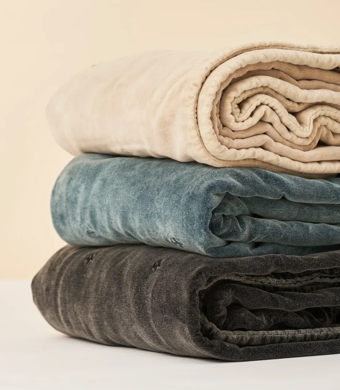 Three folded velvet blankets in cream, blue, and gray stacked on top of each other.