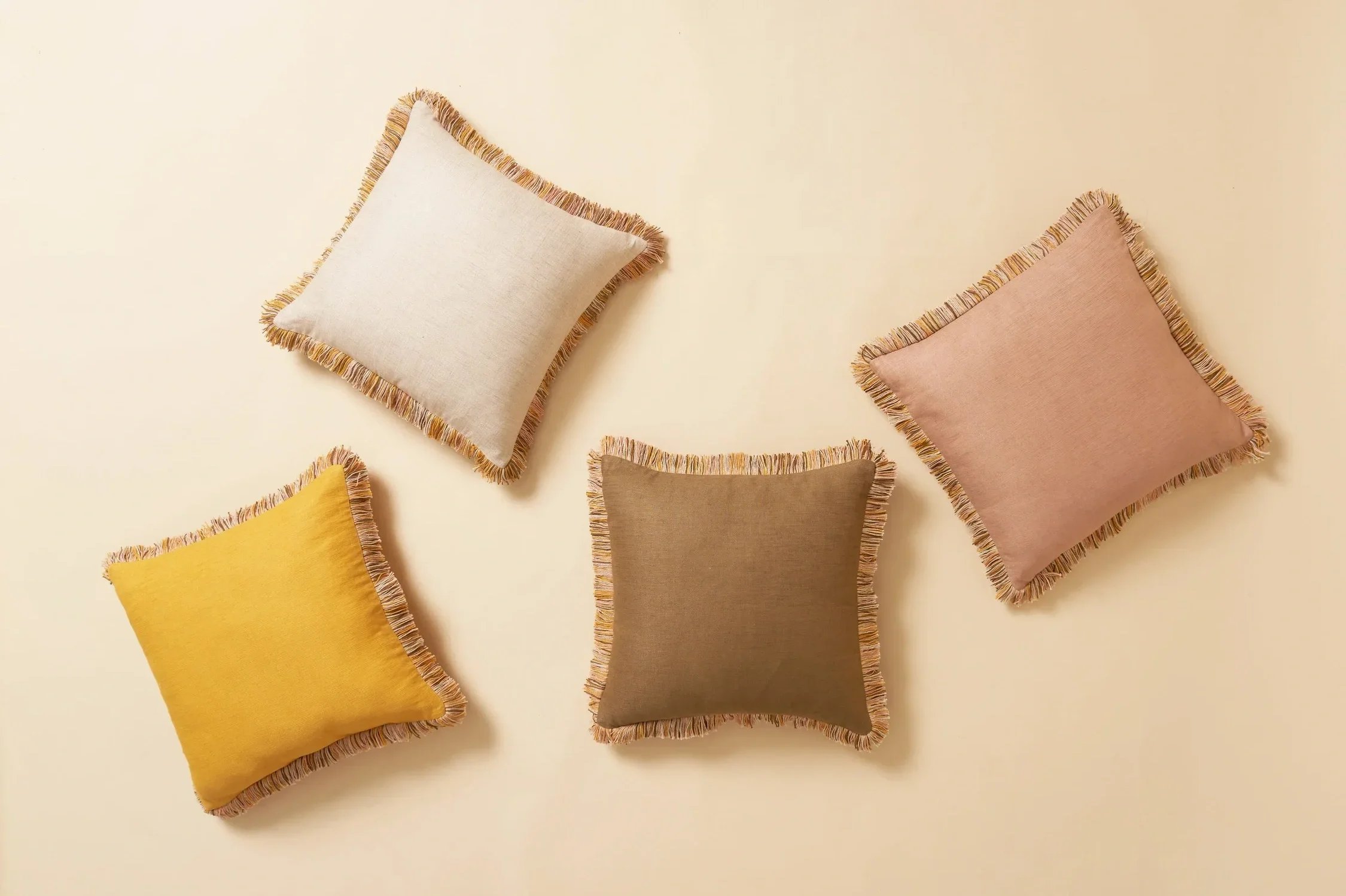 Six decorative pillows with fringed edges in shades of yellow, beige, pink, brown, and mustard on a cream-colored background.