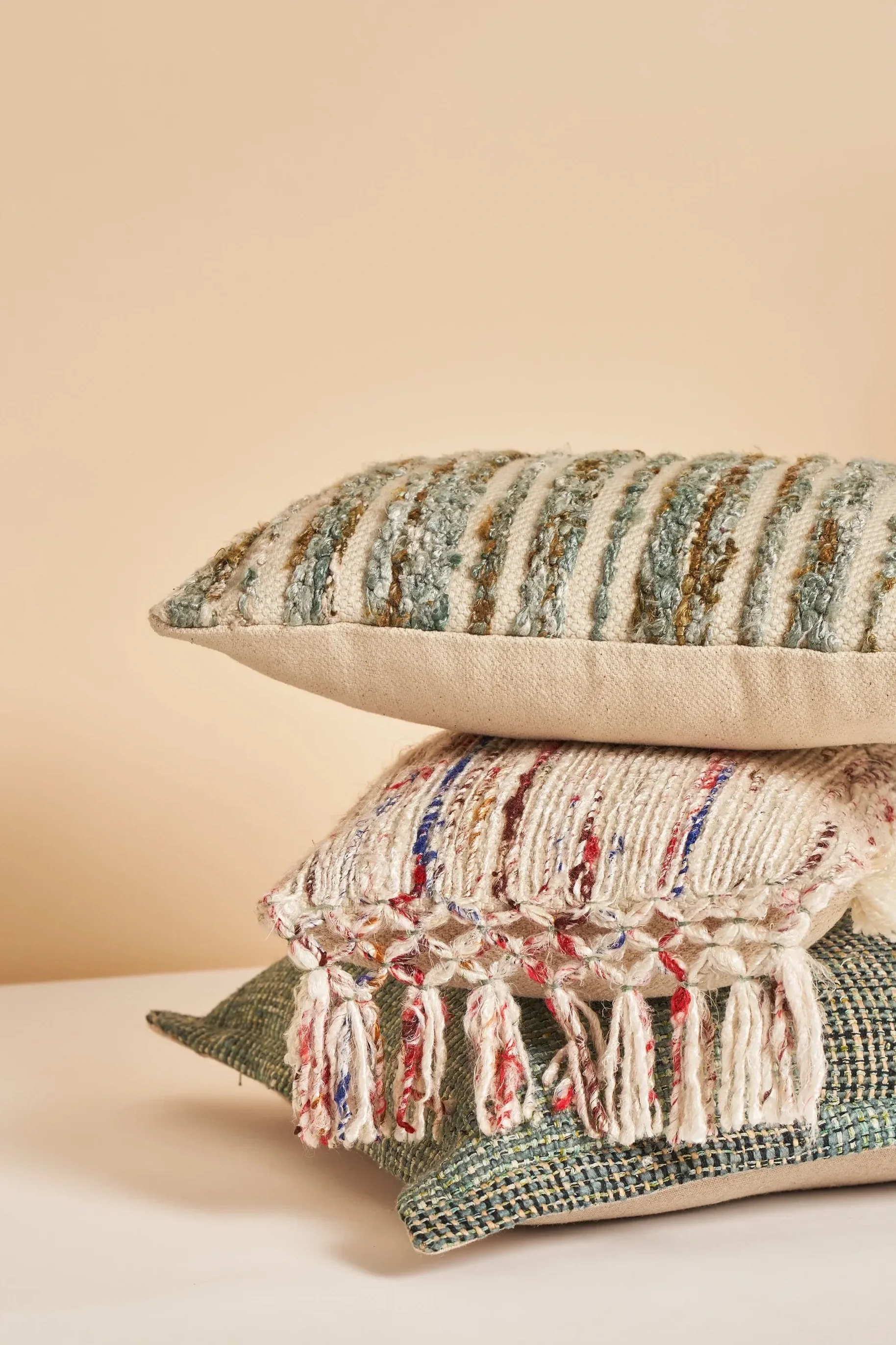 Stack of decorative pillows with various textured fabrics and colors, including beige, green, white, red, blue, and multicolored patterns.
