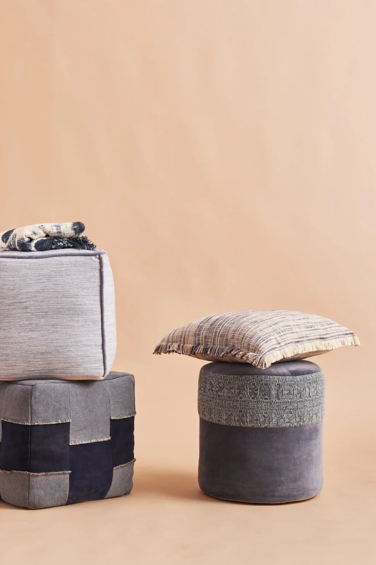 Decorative interior scene with a beige wall, a patterned fabric pillow on a cylindrical stool, and a fabric-covered storage ottoman with a blanket on top.