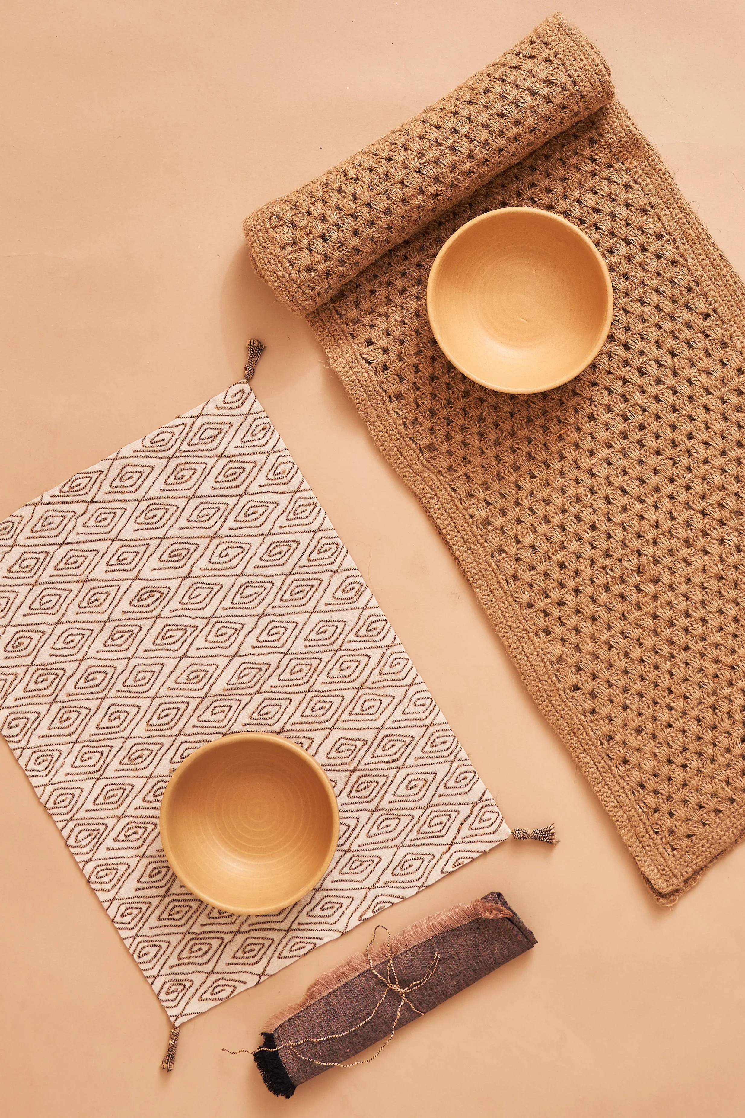 A flat lay of two woven beige table runners, one rolled up and one laid flat on a beige surface. There are two empty beige bowls, one placed on a patterned cloth with a geometric design in black and white and the other on the table runner. A small piece of dark fabric with a metallic thread and a chain is also visible.