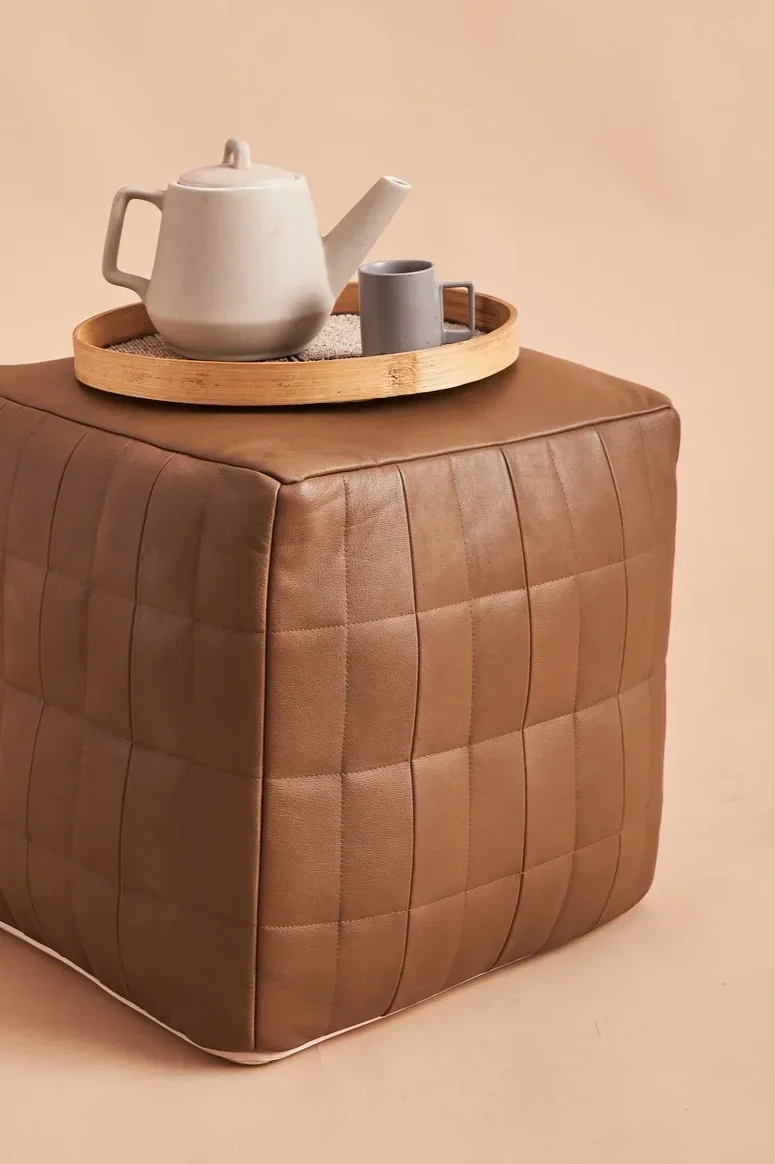 A brown quilted ottoman with a round wooden tray on top, holding a white teapot and a gray mug, against a beige background.