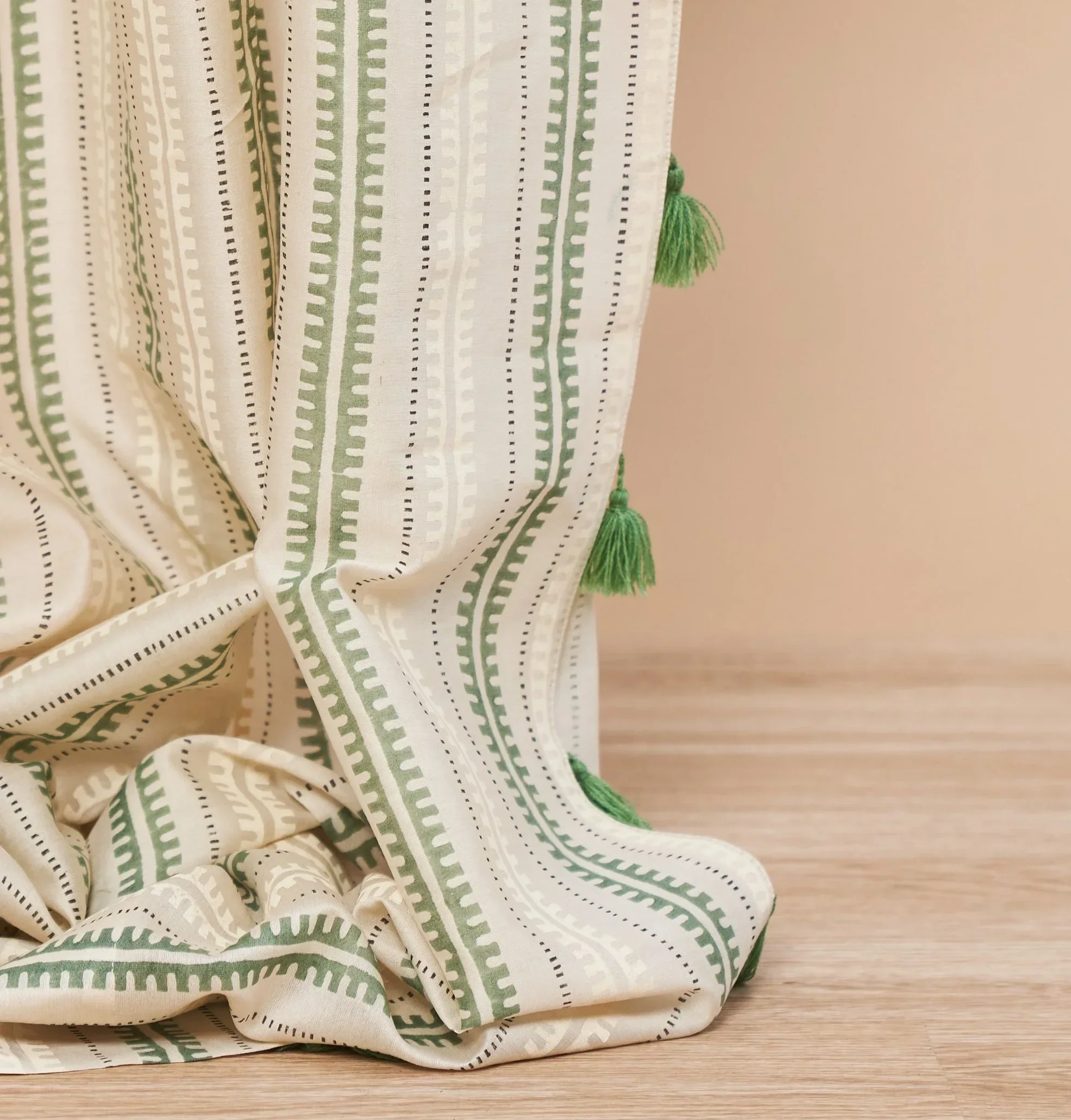 Close-up of a curtain with green and white patterned fabric and green tassels, hanging near a wooden surface.