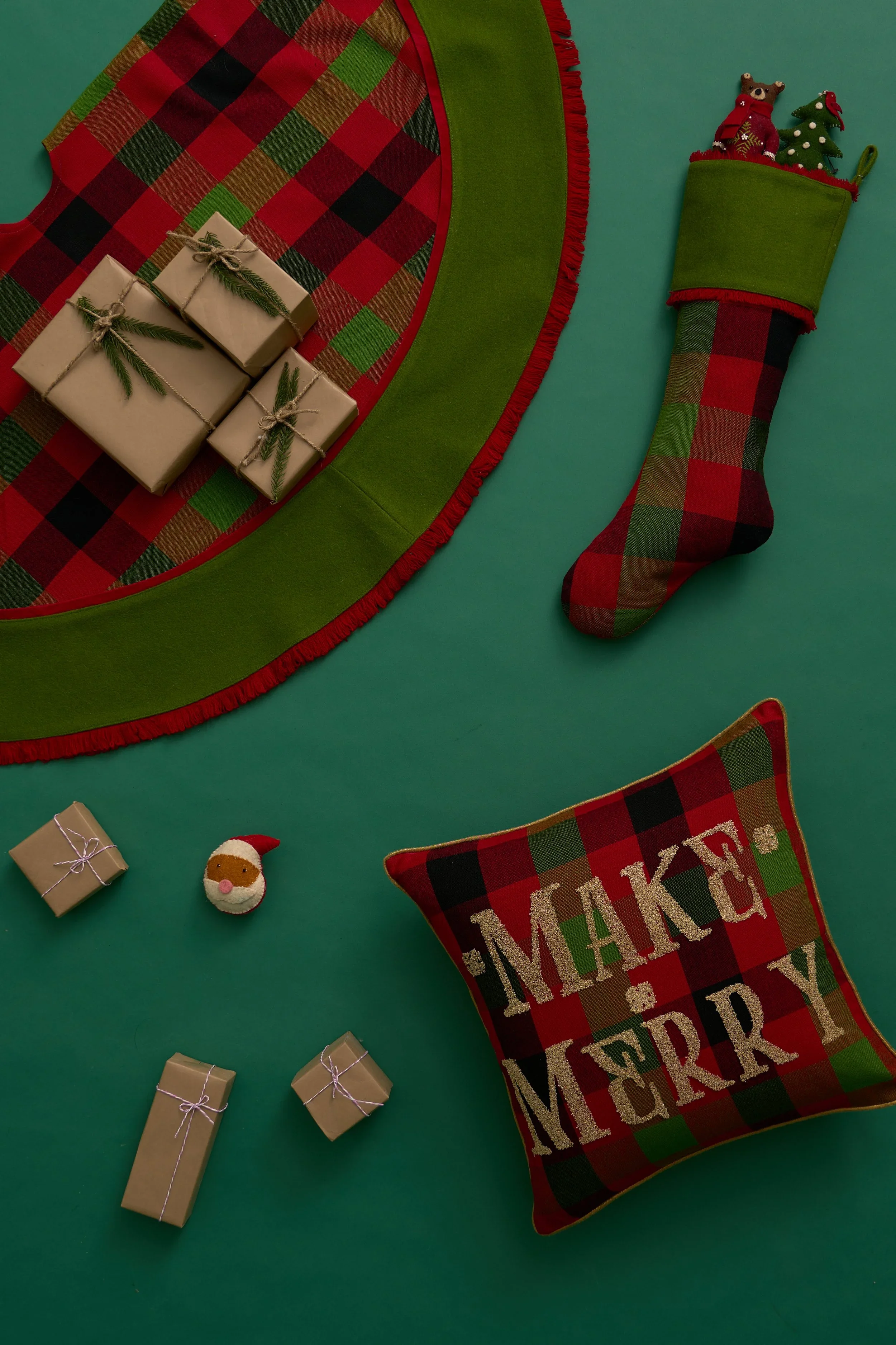 Green background with Christmas decorations including a red and green plaid stocking holding small plush reindeer and Christmas tree, a green and plaid Christmas pillow with "MAKE A MERRY" written on it, small wrapped gifts with ribbons, and a Santa face ornament.