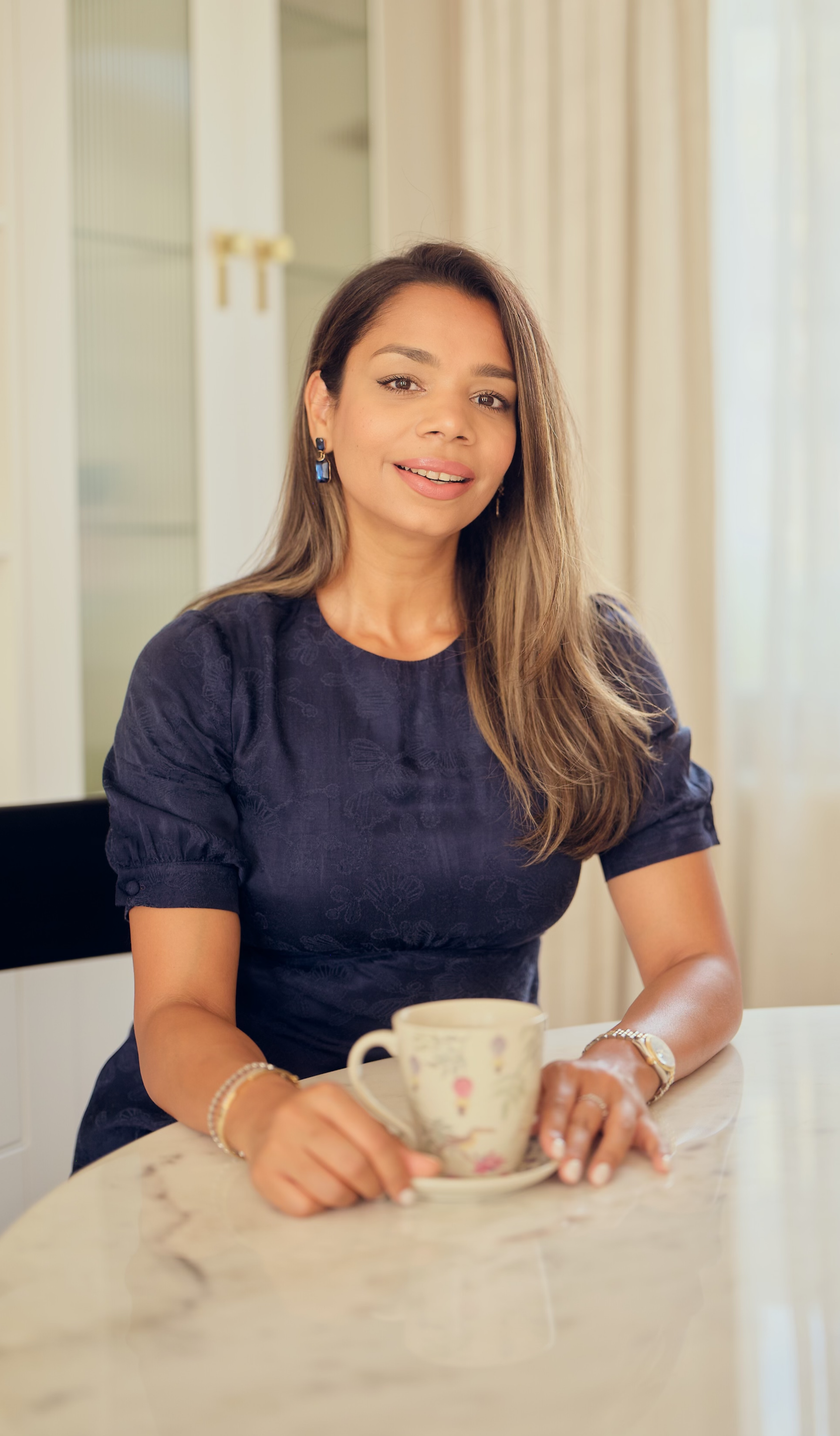 A woman with brown hair and earrings sitting at a table, holding a floral teacup and saucer, in a well-lit room with curtains.