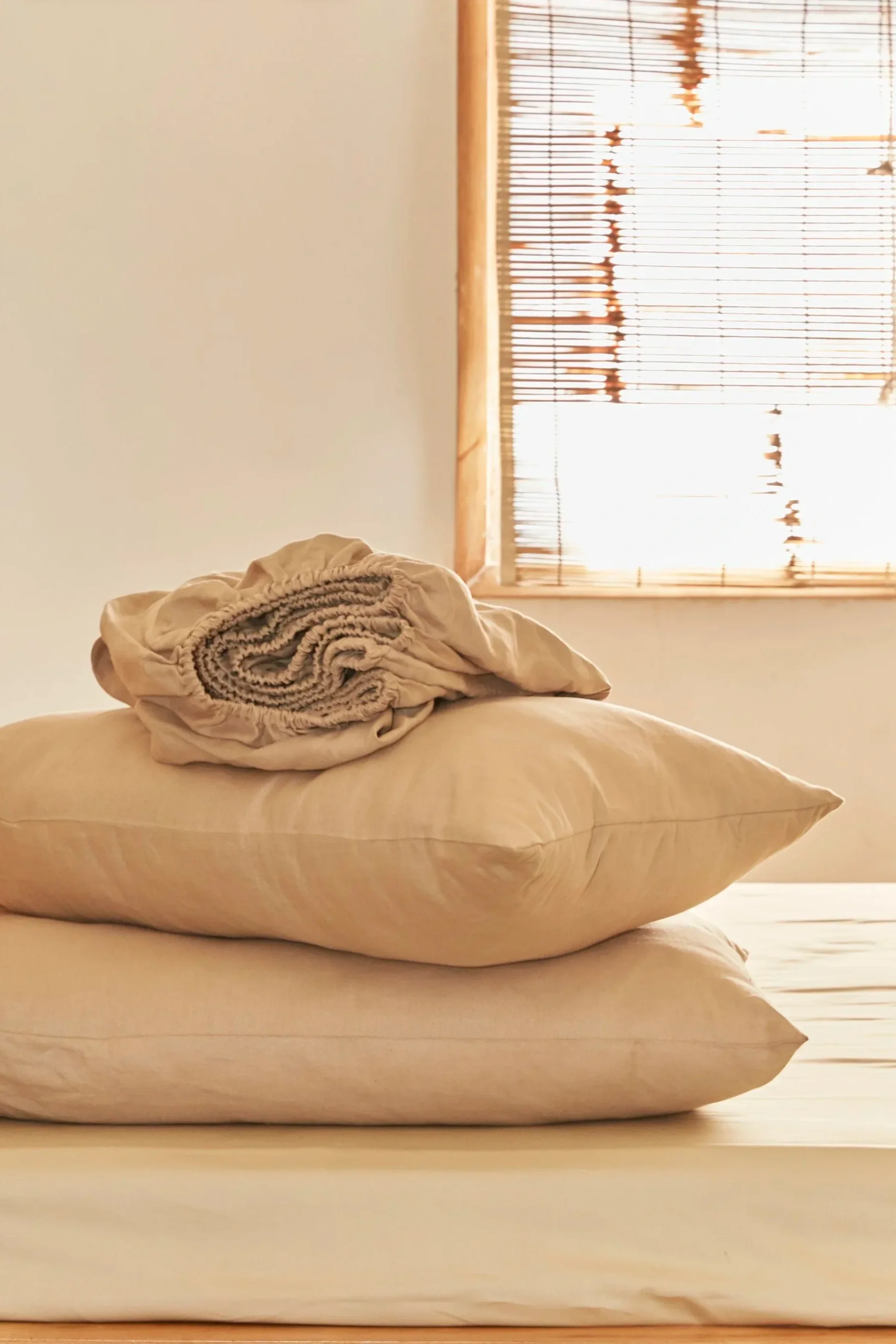 A bedroom scene with two beige pillows stacked on top of each other, a rolled-up blanket on the upper pillow, and a window with horizontal blinds letting in sunlight in the background.
