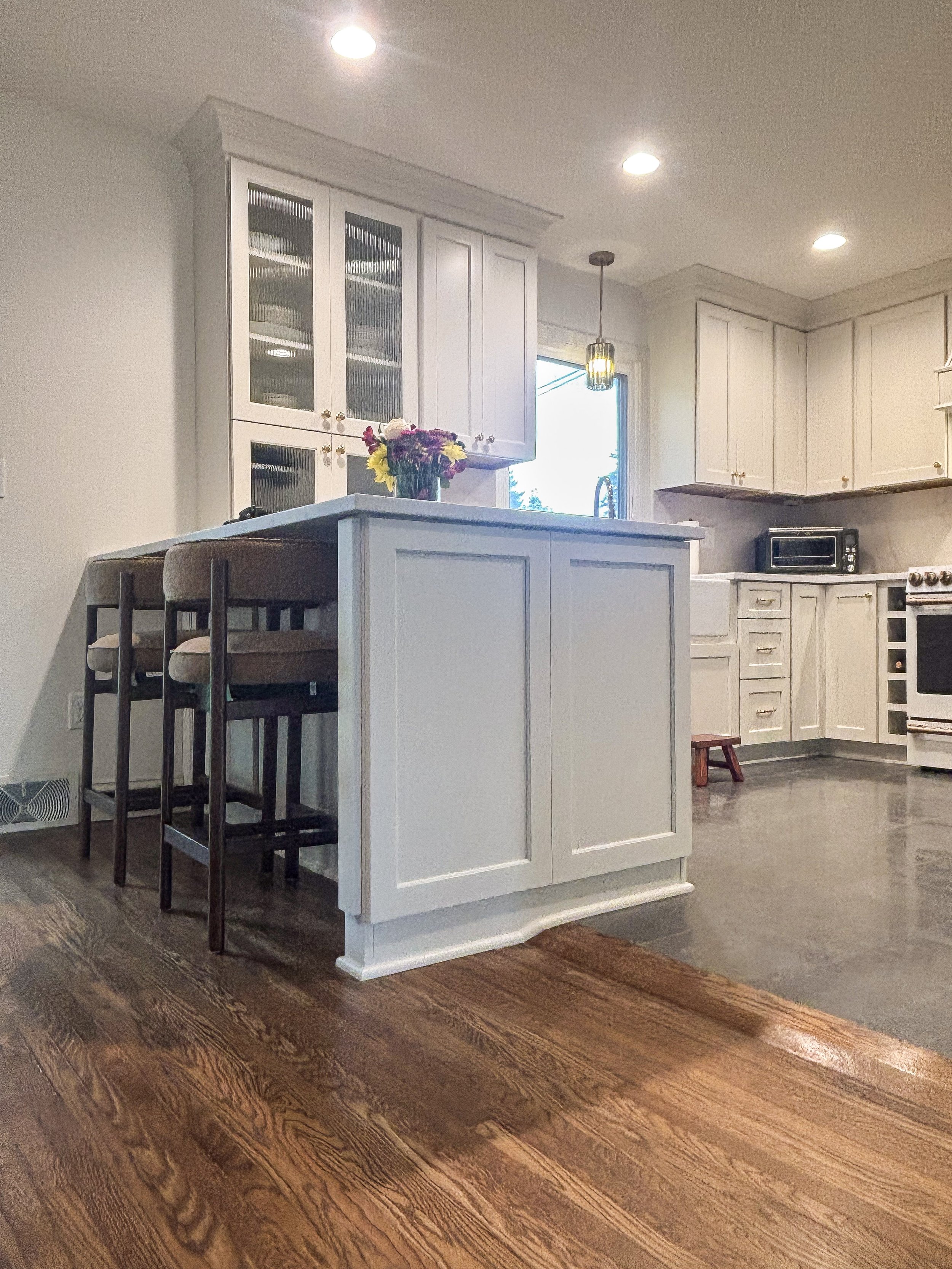 A kitchen with white cabinets, a white island, a black microwave, and a wooden floor. There is a vase with colorful flowers on the island, two chairs at the island, and a window with a hanging pendant light.