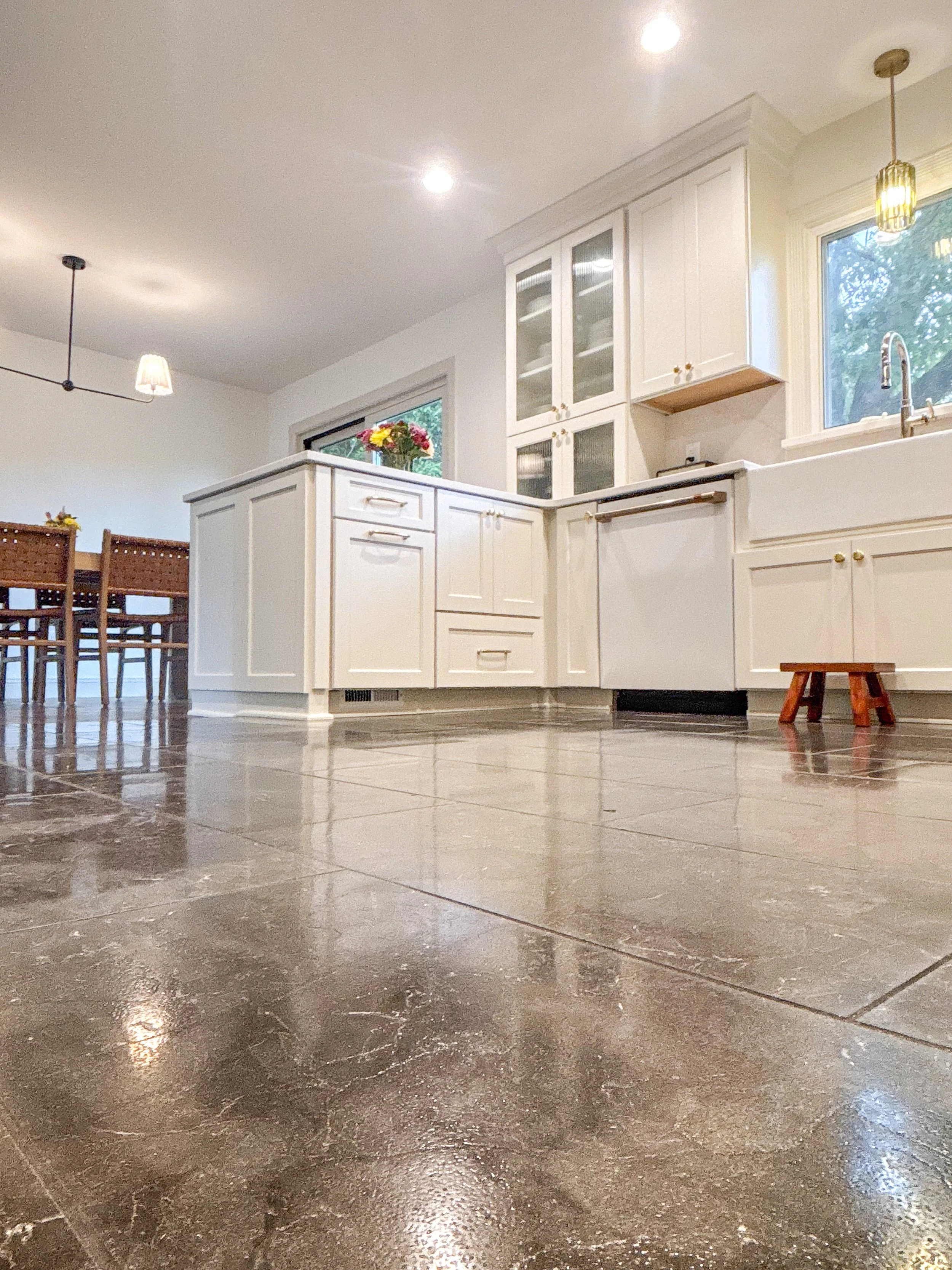 View of a kitchen with white cabinets and large windows, a tiled floor, a small wooden stool, and a dining table with chairs in the background.