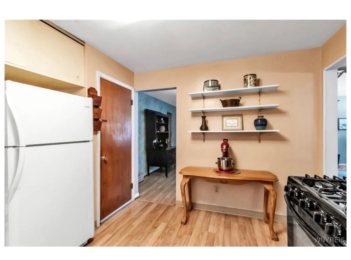 Kitchen with wooden floor, white refrigerator on the left, wooden door in the center, beige walls, wooden table with a red stand mixer, white shelves with decorative items above the table, and a stove on the right.