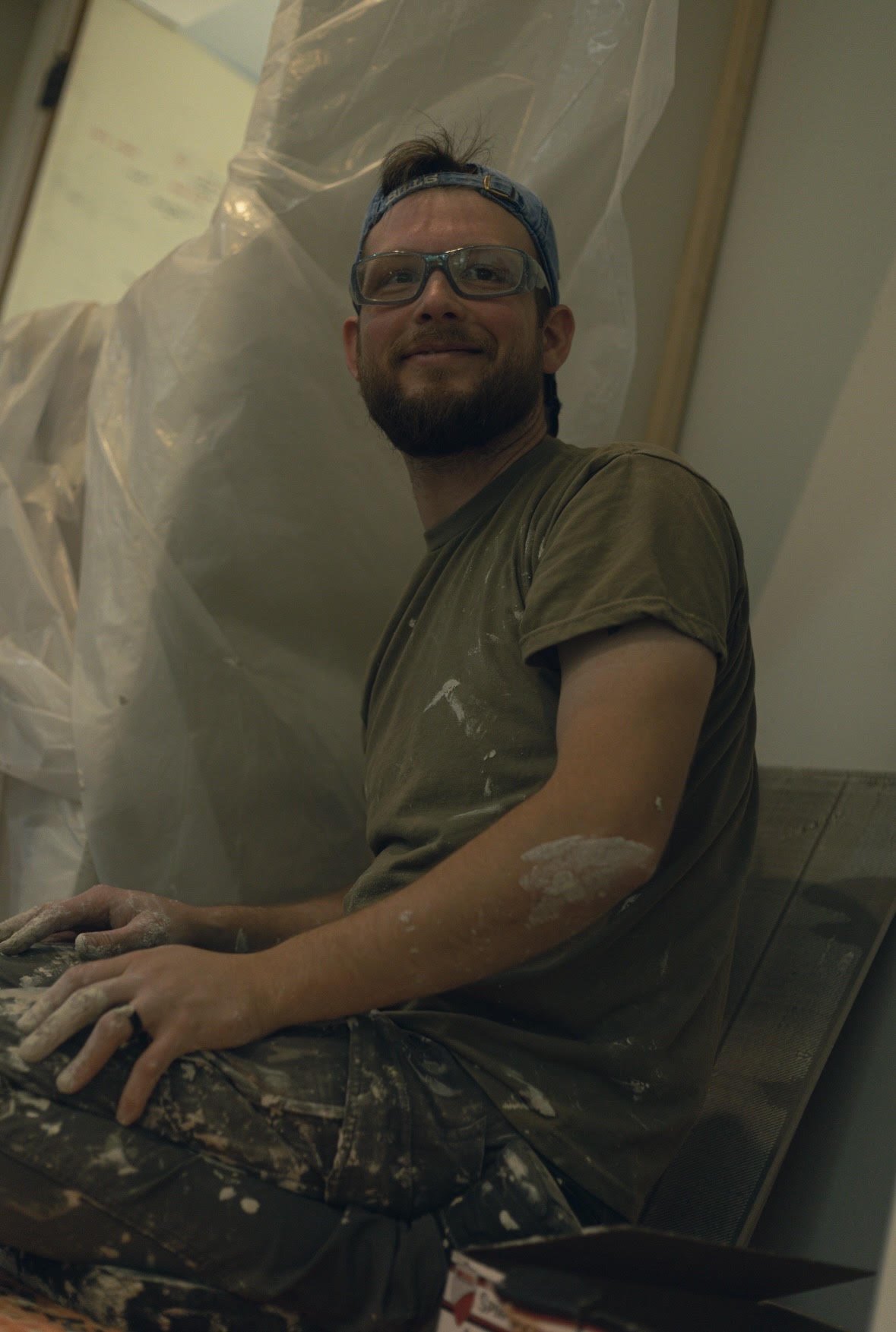 A man with glasses, a cap backward, and a beard, smiling while working on a craft project, wearing a paint-stained shirt and camouflage pants, inside a room with a wall and a plastic sheet in the background.