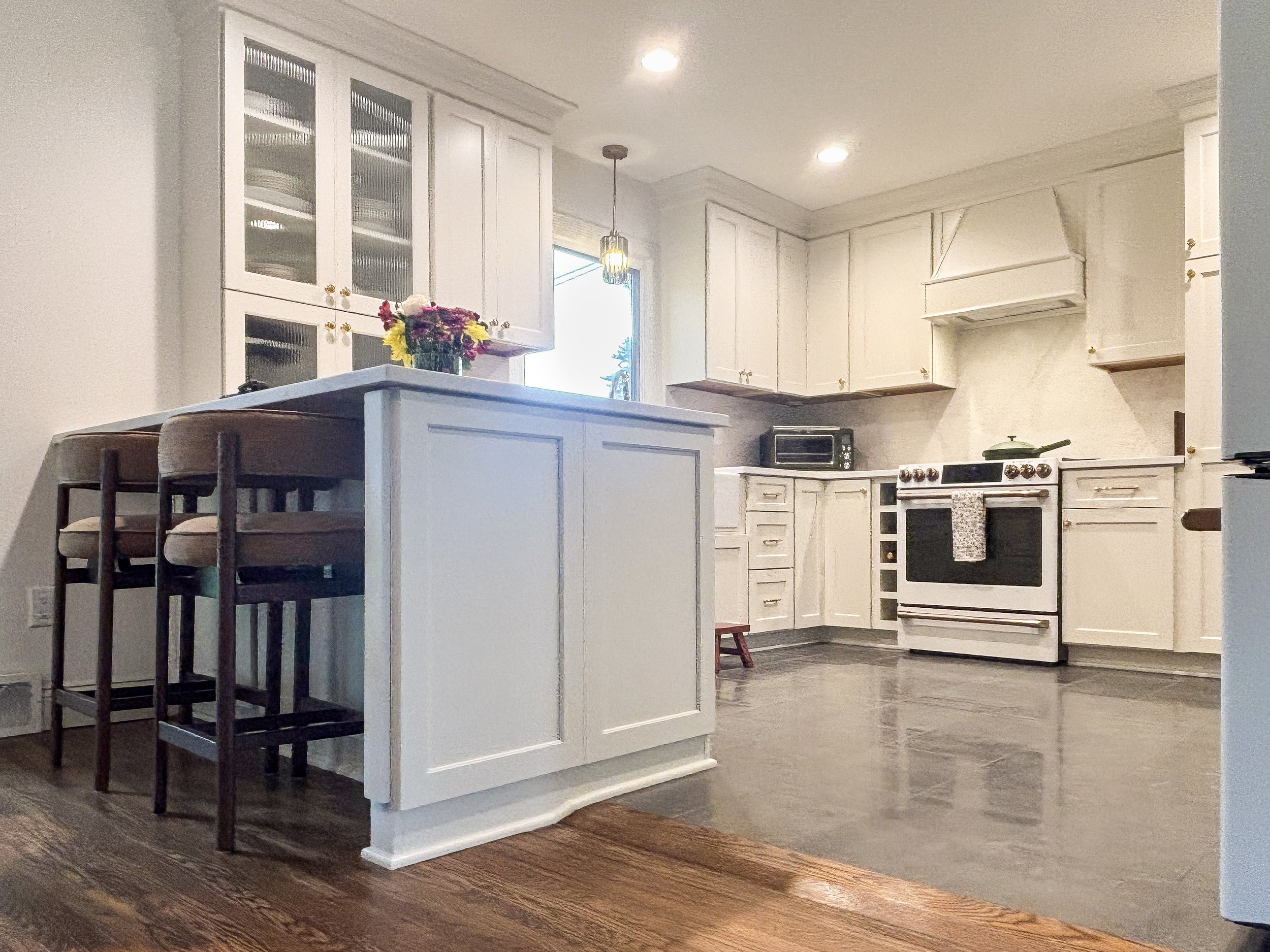 A kitchen with white cabinets, a white stove, a black microwave, wooden stools at a kitchen island, a vase of colorful flowers, and a window with a view of trees outside.