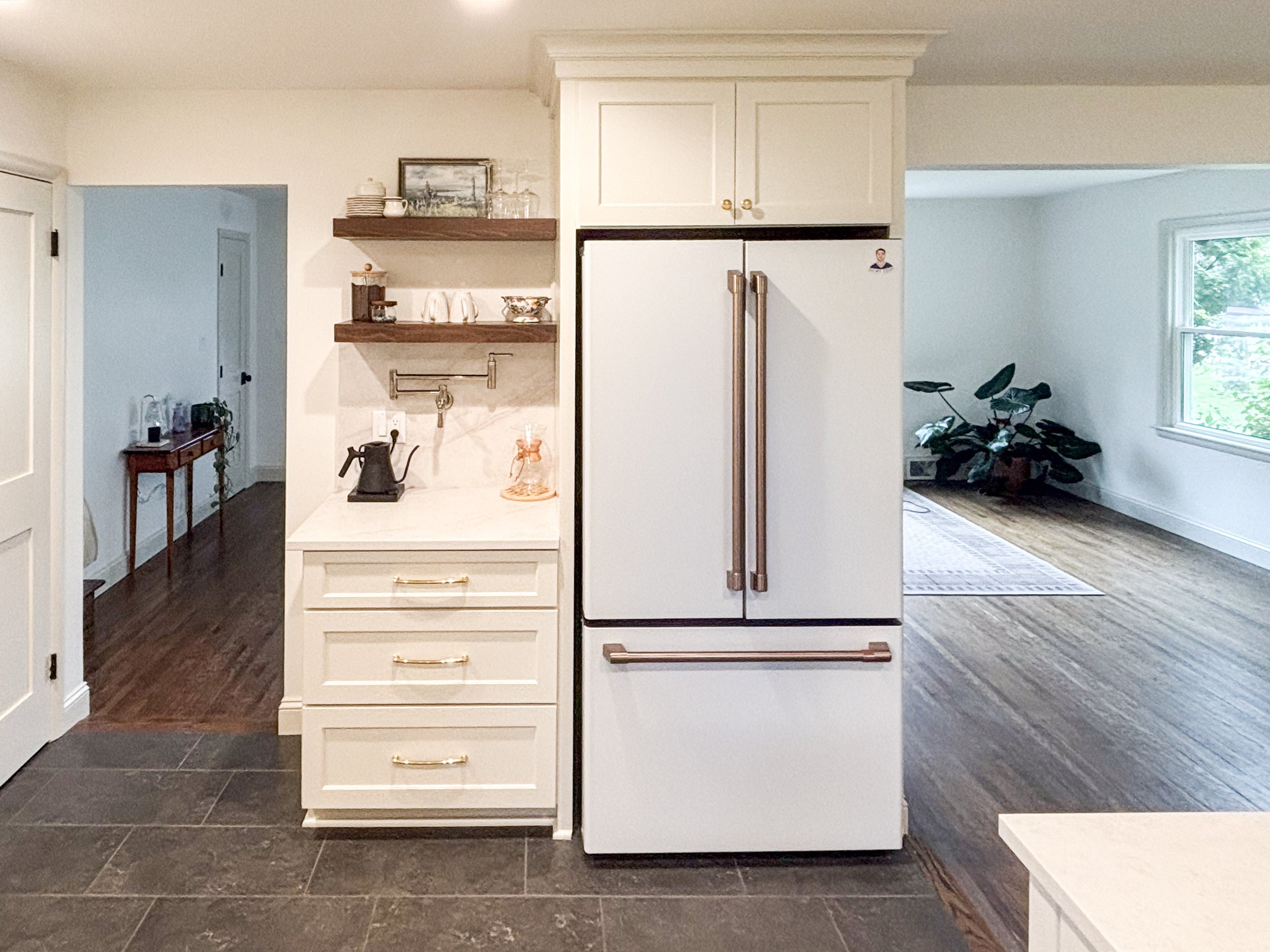 A kitchen with white cabinets, a double-door refrigerator, floating wooden shelves with dishes, glasses, and a painting, and dark tiled flooring. Adjacent to the kitchen is a living room with a window, a potted plant, and an exercise bike.