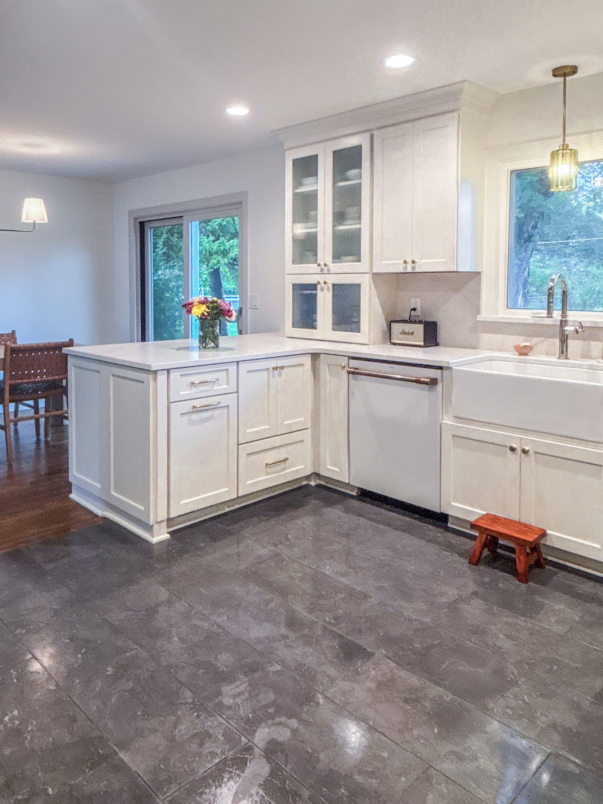 A kitchen with white cabinets, a white island with a bouquet of flowers, and black flooring. There is a window over the sink, a small wooden step stool, and a view of trees outside.