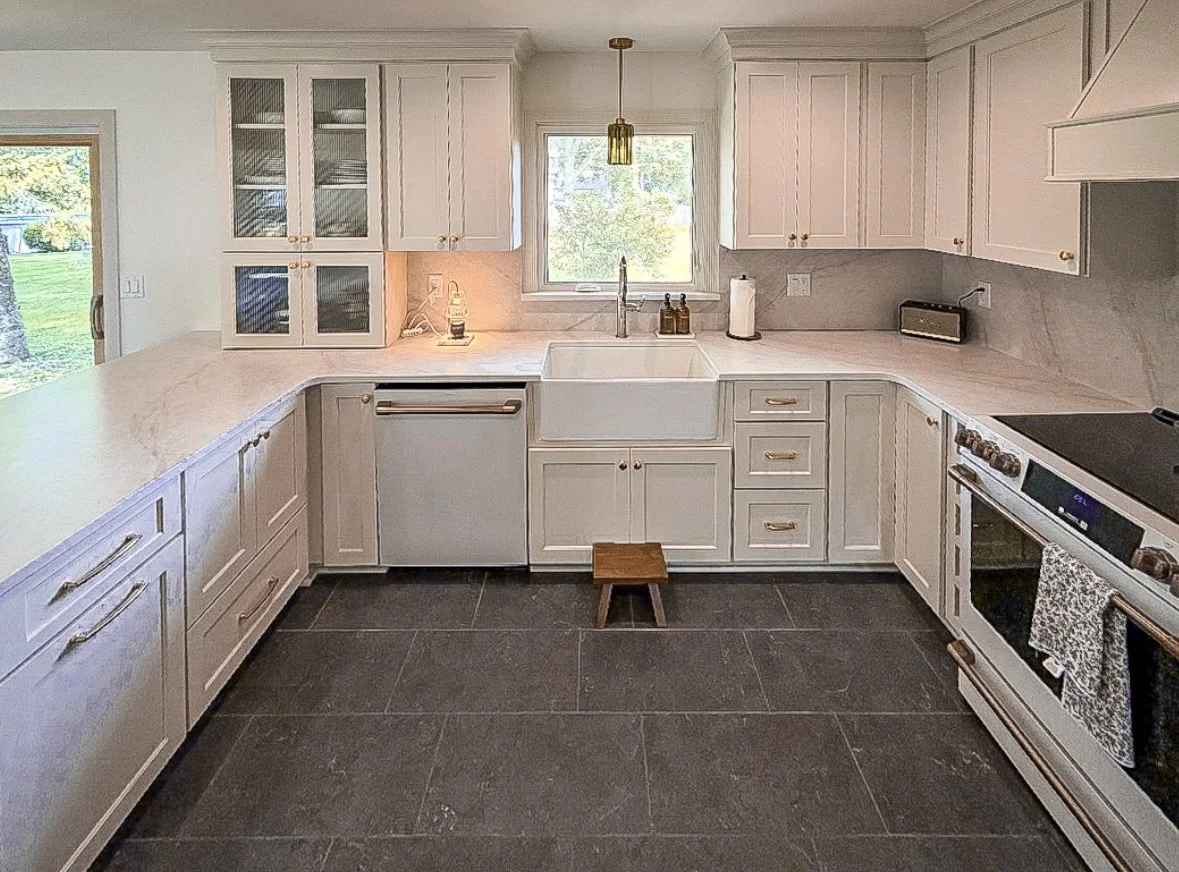 White kitchen with cabinets, a farmhouse sink, a window above the sink, and a stove with an oven. The countertop has a small stool, a paper towel roll, and a small radio.