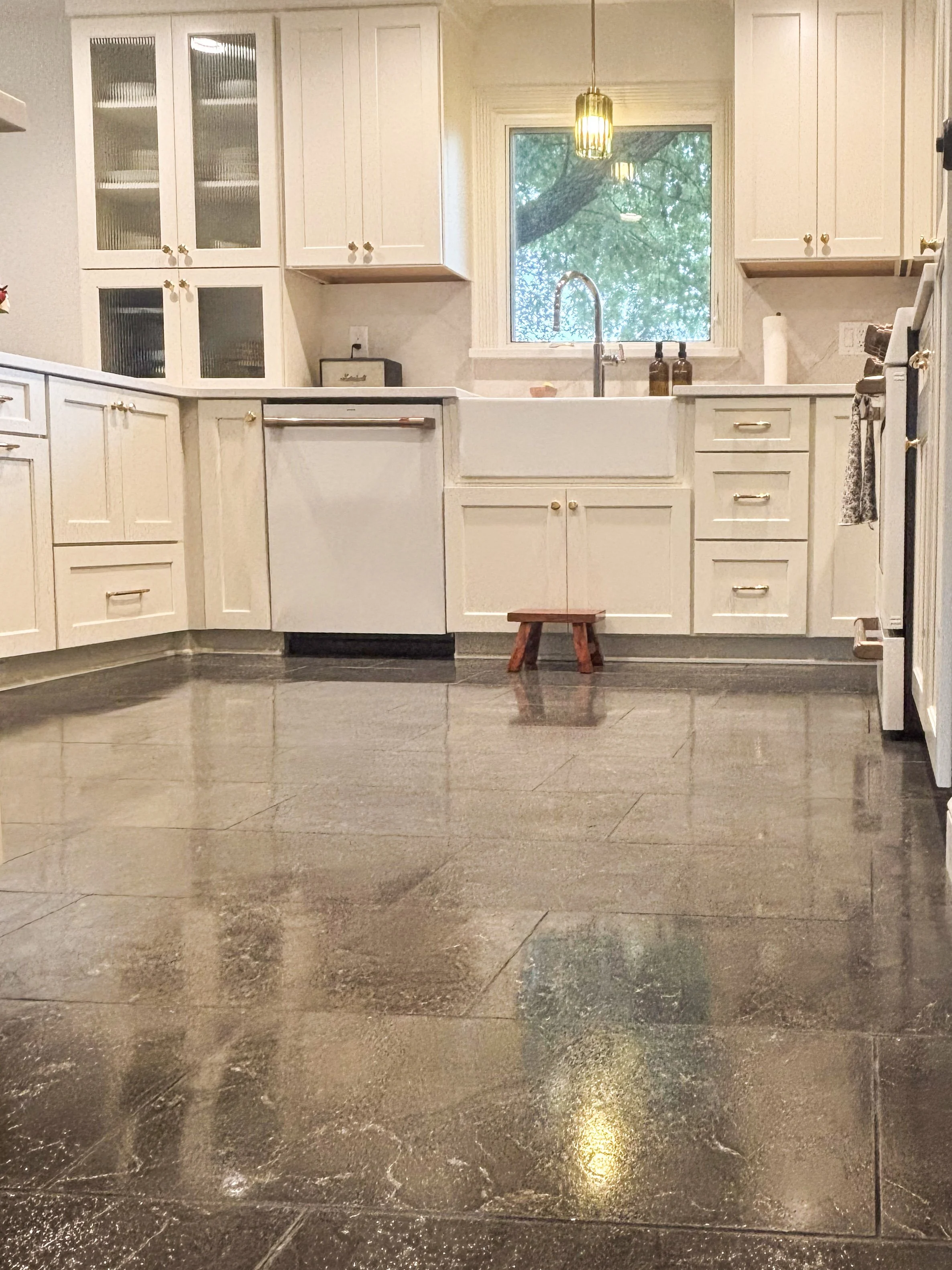 A kitchen with white cabinets, a large window, a sink under the window, and two brown soap dispensers. There is a gold pendant light hanging from the ceiling and a small wooden stool in front of the sink. The floor is reflective and dark-colored.
