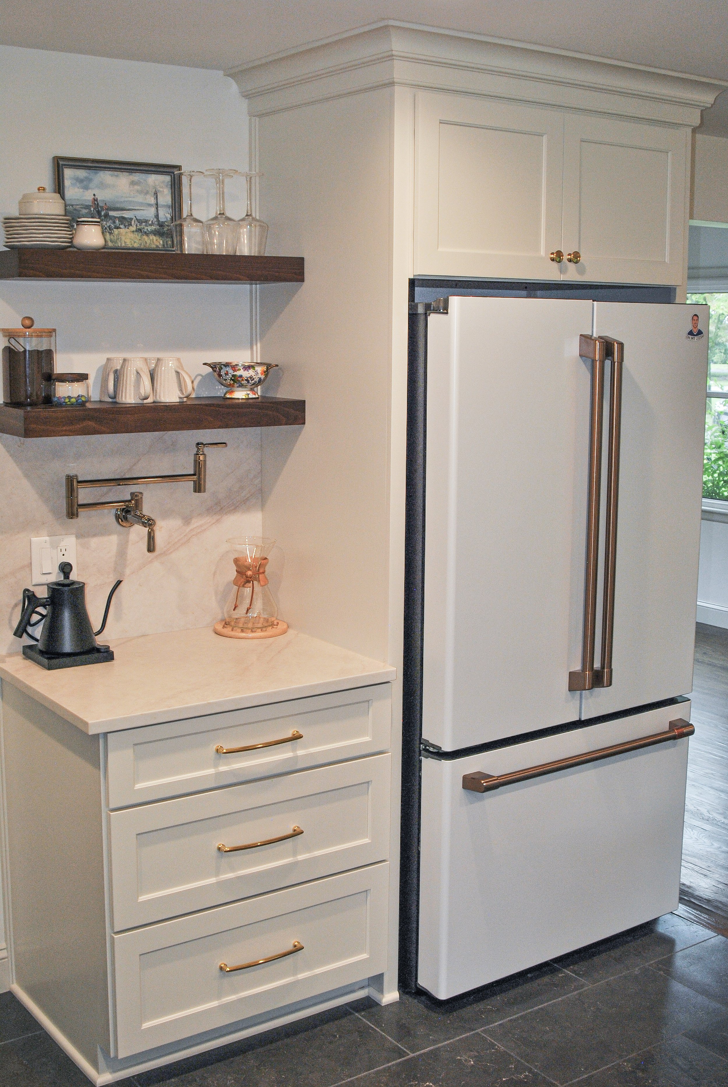 Kitchen corner with a white chest of drawers, a white vintage refrigerator with bronze handles, and dark stone tiles on the floor. Open shelves above display glassware, dishes, and a landscape painting.